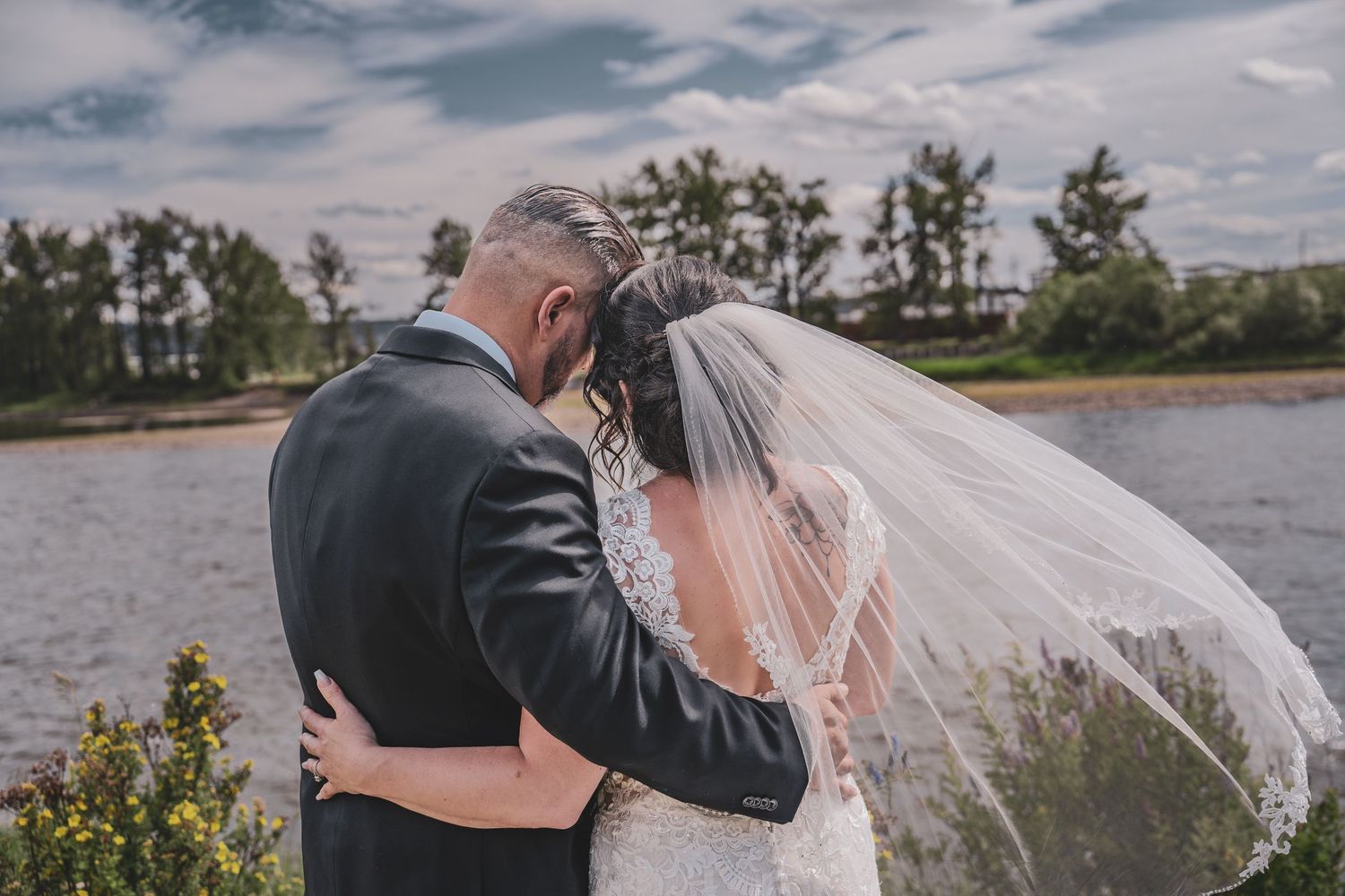 Bride and groom sharing a romantic embrace by a lakeside with trees and cloudy sky behind them.