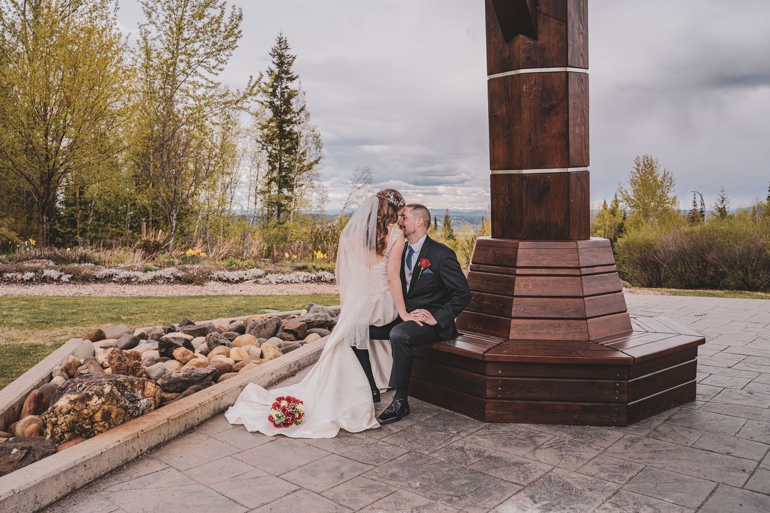 Bride and groom sitting closely together beside a tall stone monument in a garden setting.
