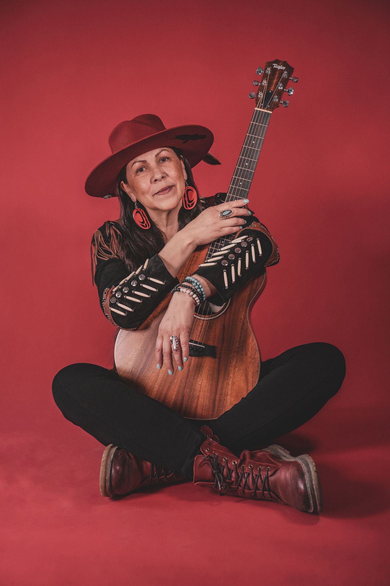 Kym Gouchie in red hat holding acoustic guitar seated cross-legged on red studio floor during musician photoshoot.
