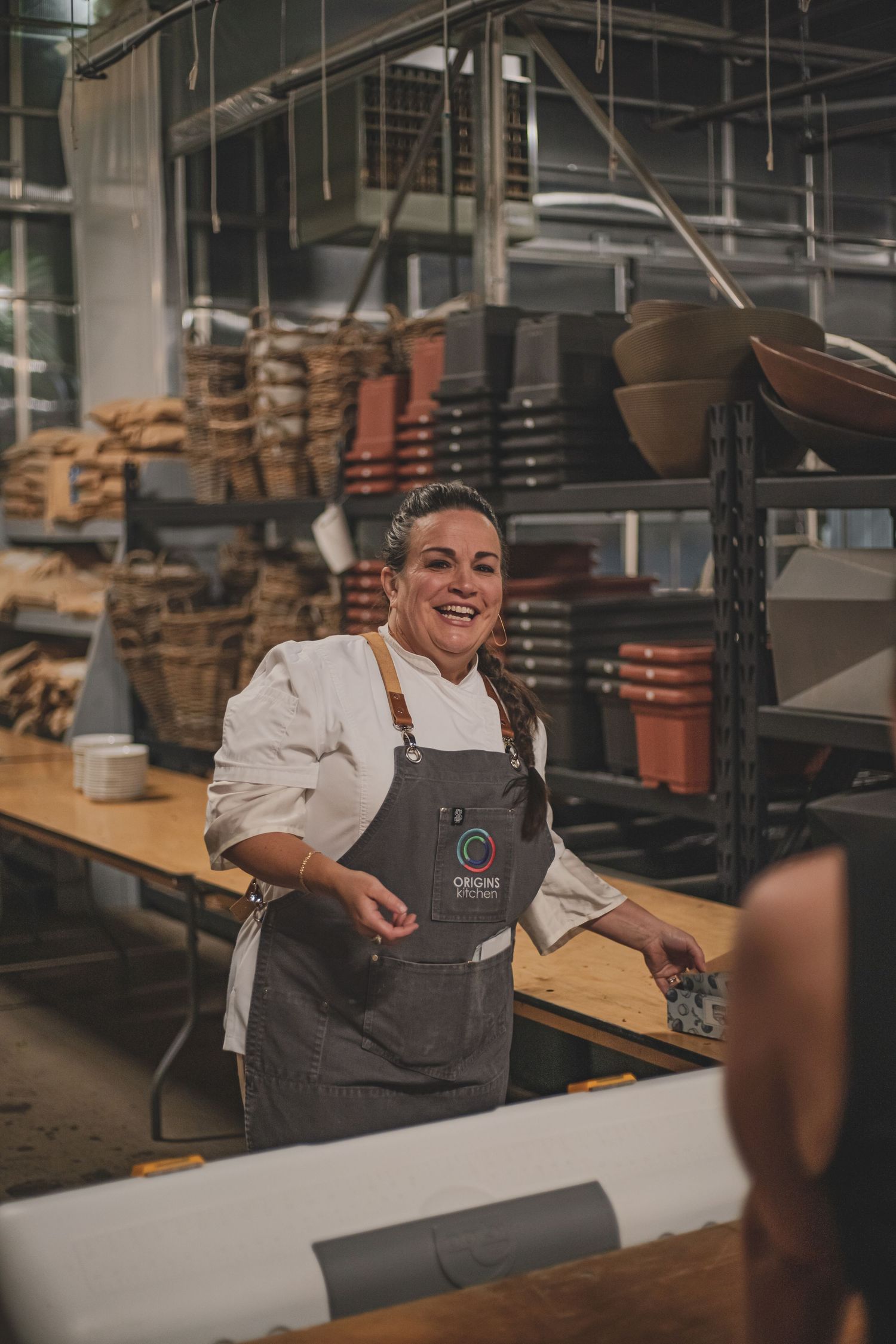 A smiling female chef in a white coat and gray apron stands in a rustic kitchen storage area.