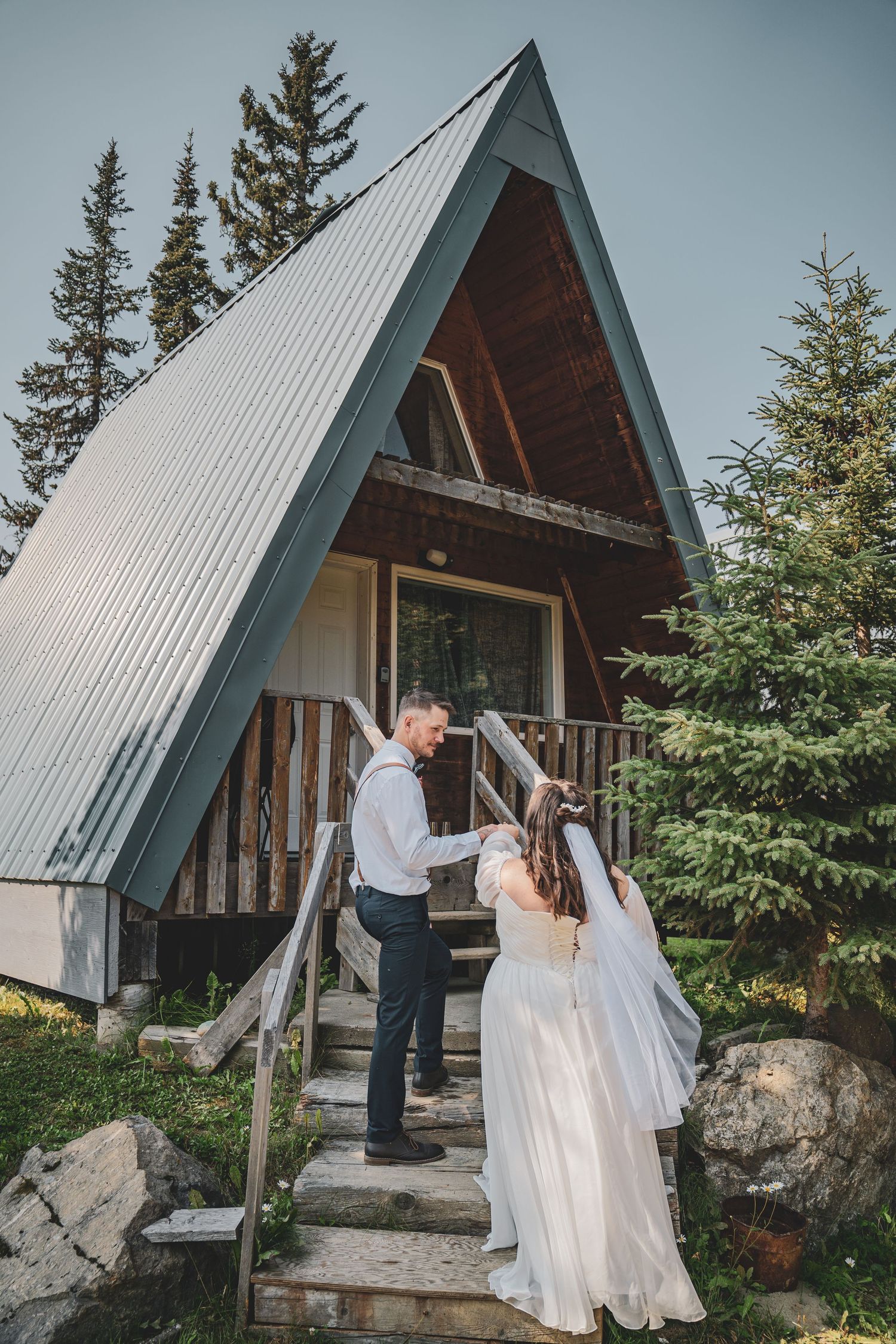 Bride and groom walk up stone steps to a charming A-frame cabin surrounded by evergreen trees at Azouzetta Lake Lodge elopement.