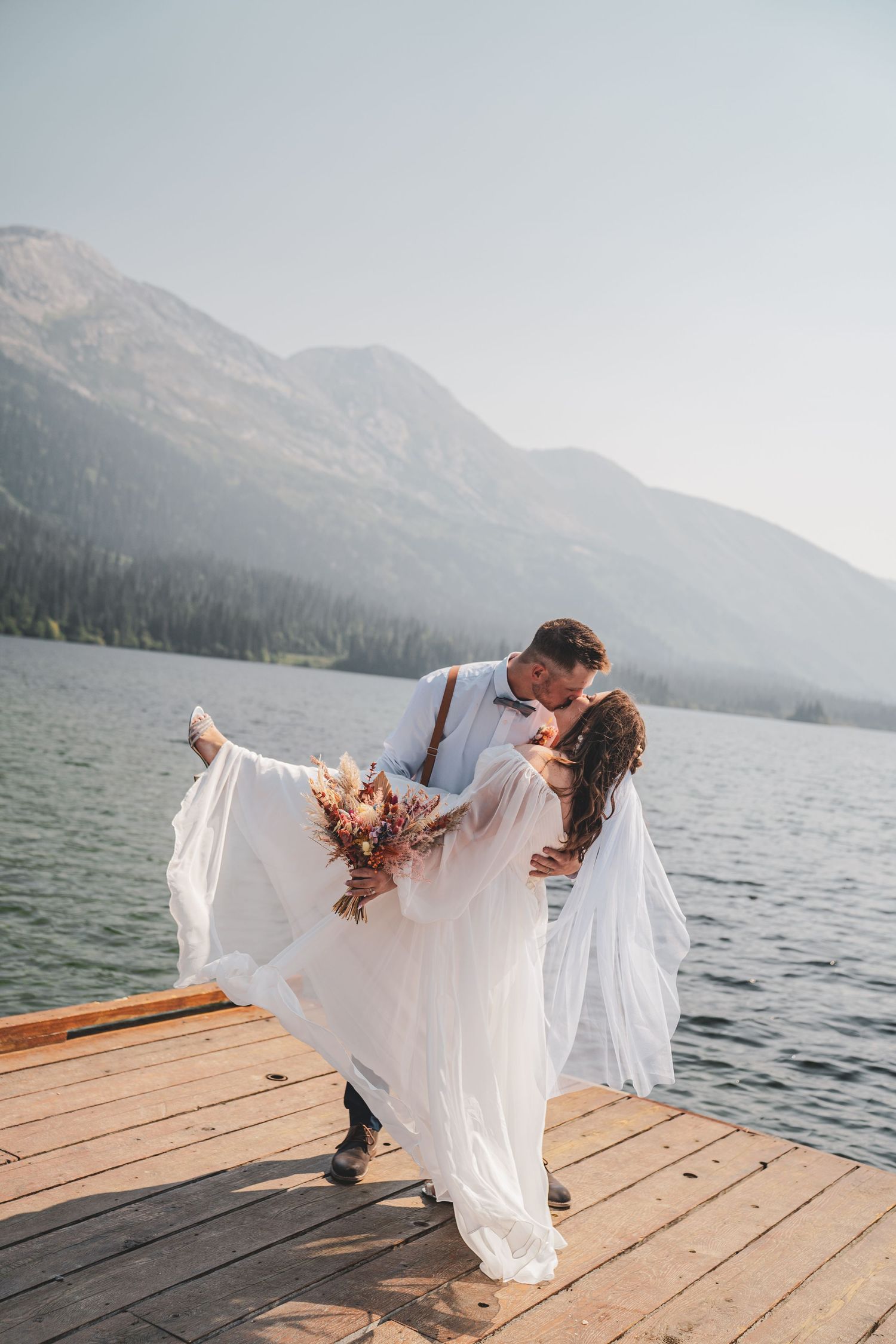 Groom dips bride in white dress on wooden dock with stunning mountain lake view during their elopement.
