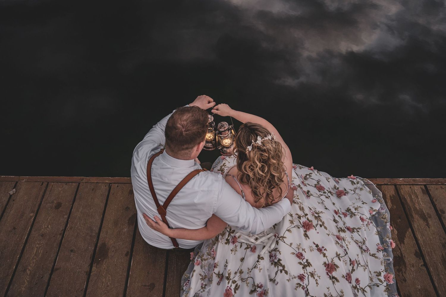 Bride and groom sit together on wooden dock overlooking dramatic dark cloudy lake at their elopement.