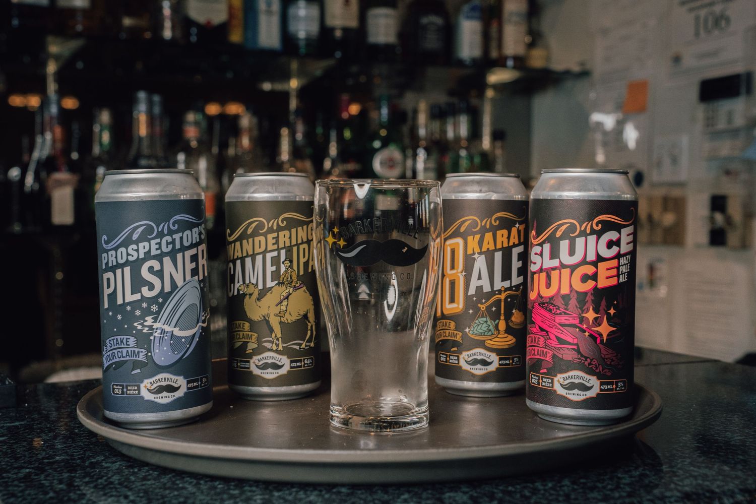 Four craft beer cans including Prospector Pilsner and Sluice Juice displayed on a bar tray.