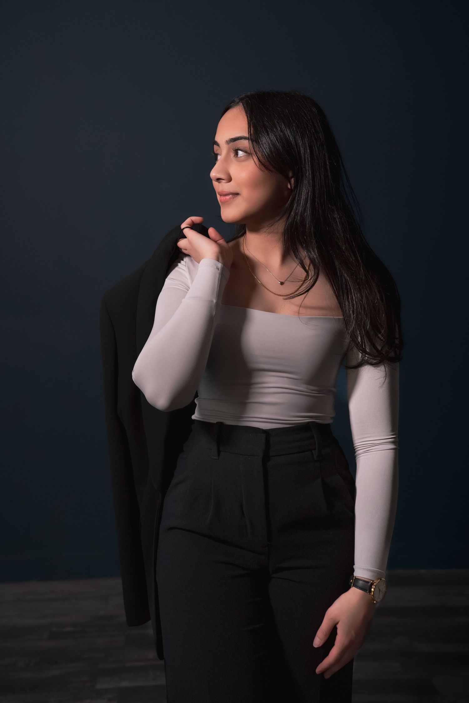 Young woman in white top holding black blazer over shoulder against dark background during her headshot photoshoot.
