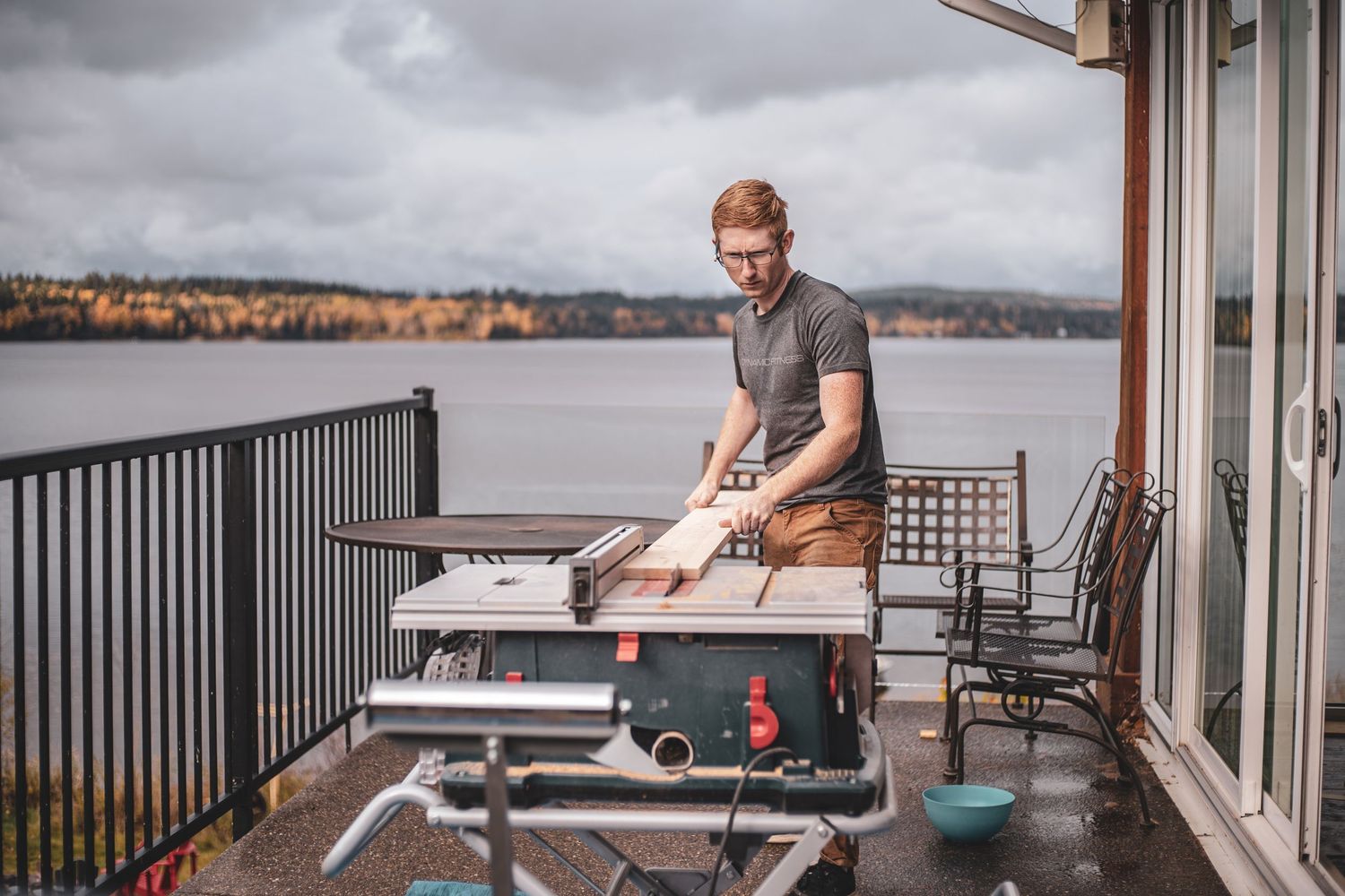 Red-haired carpenter using table saw on balcony overlooking scenic lake on overcast day.