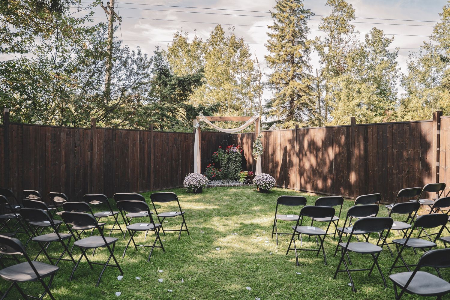Outdoor backyard wedding ceremony setup with black folding chairs and wooden fence backdrop.