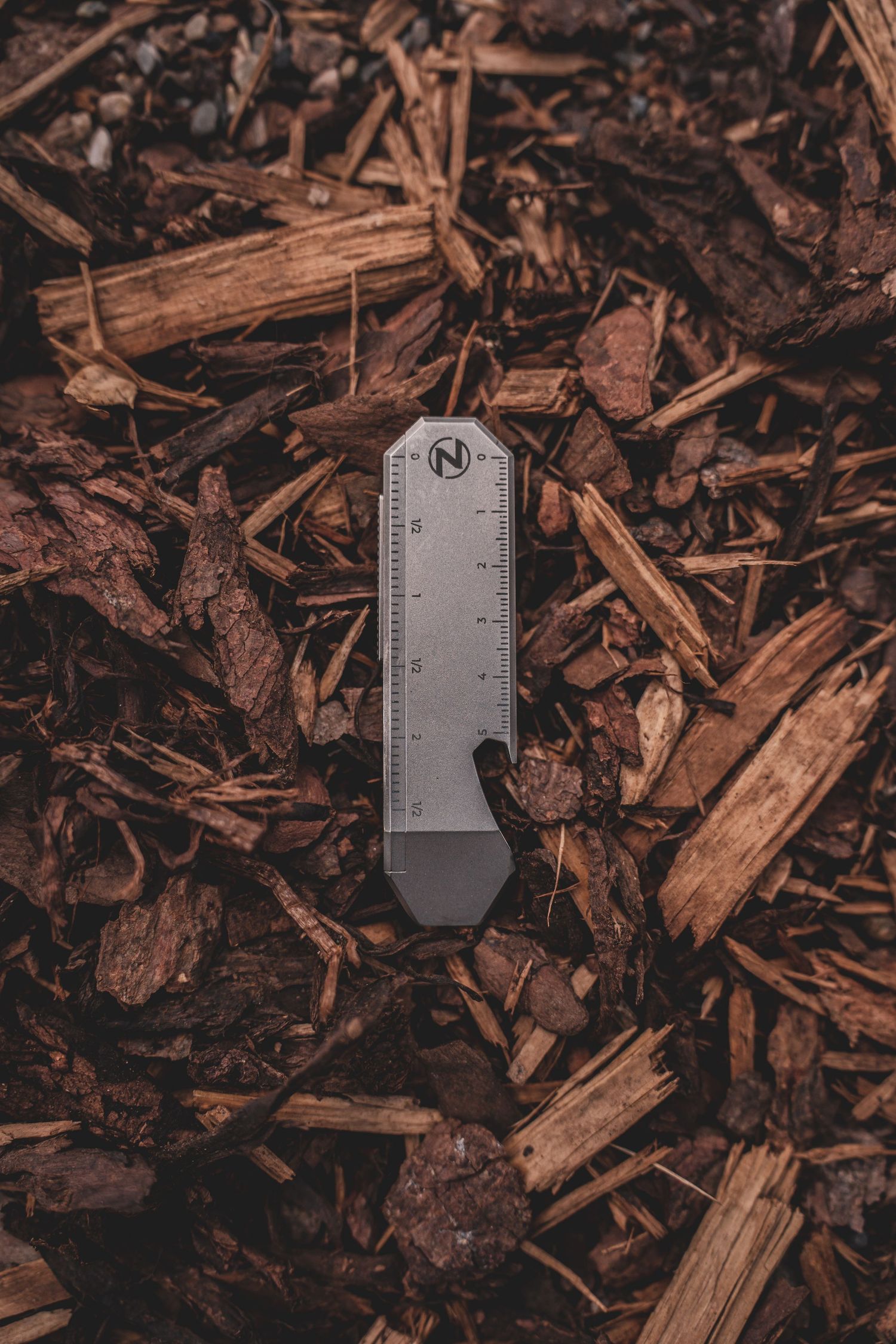 Product photography of Minimalist gray multi-tool ruler lying on rustic brown wood chip mulch ground.
