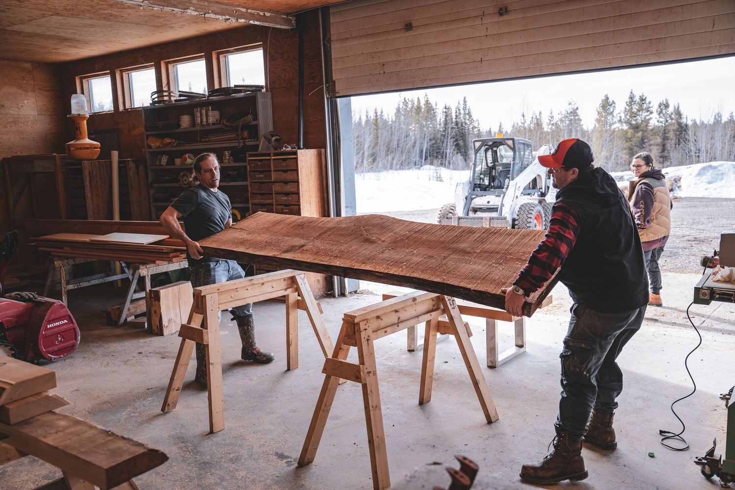 Workers handling a large wooden slab on sawhorses inside a workshop with snowy landscape outside.