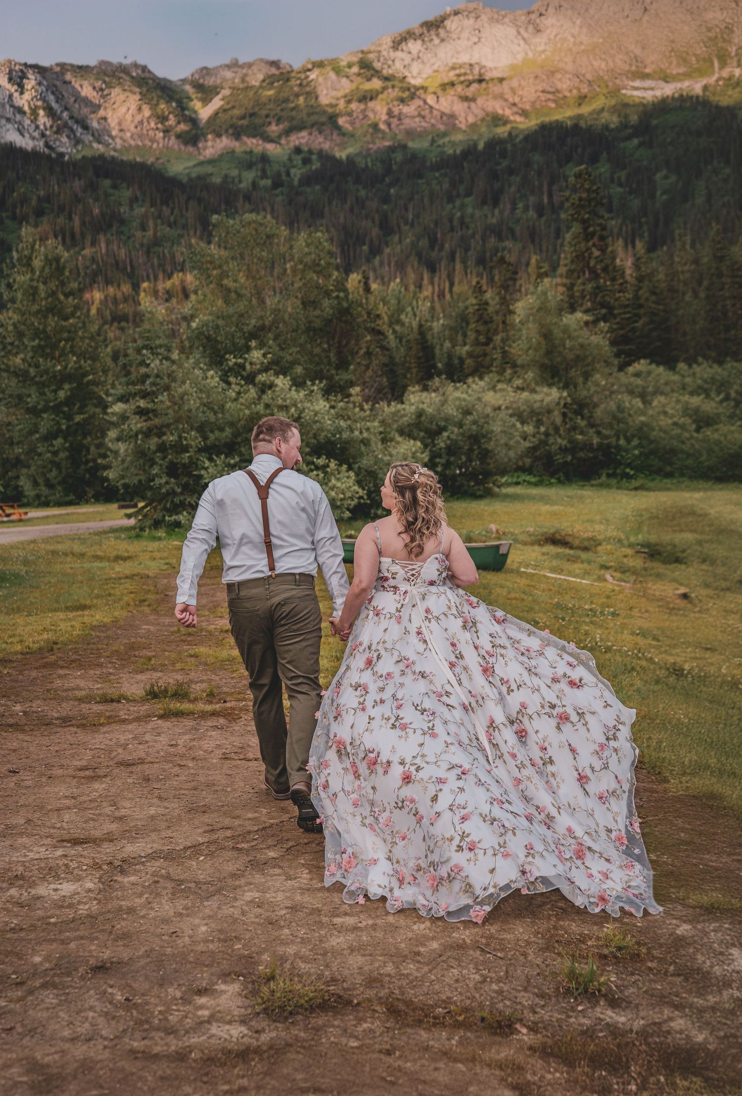 Bride in floral gown and groom walking hand in hand toward scenic mountain landscape.