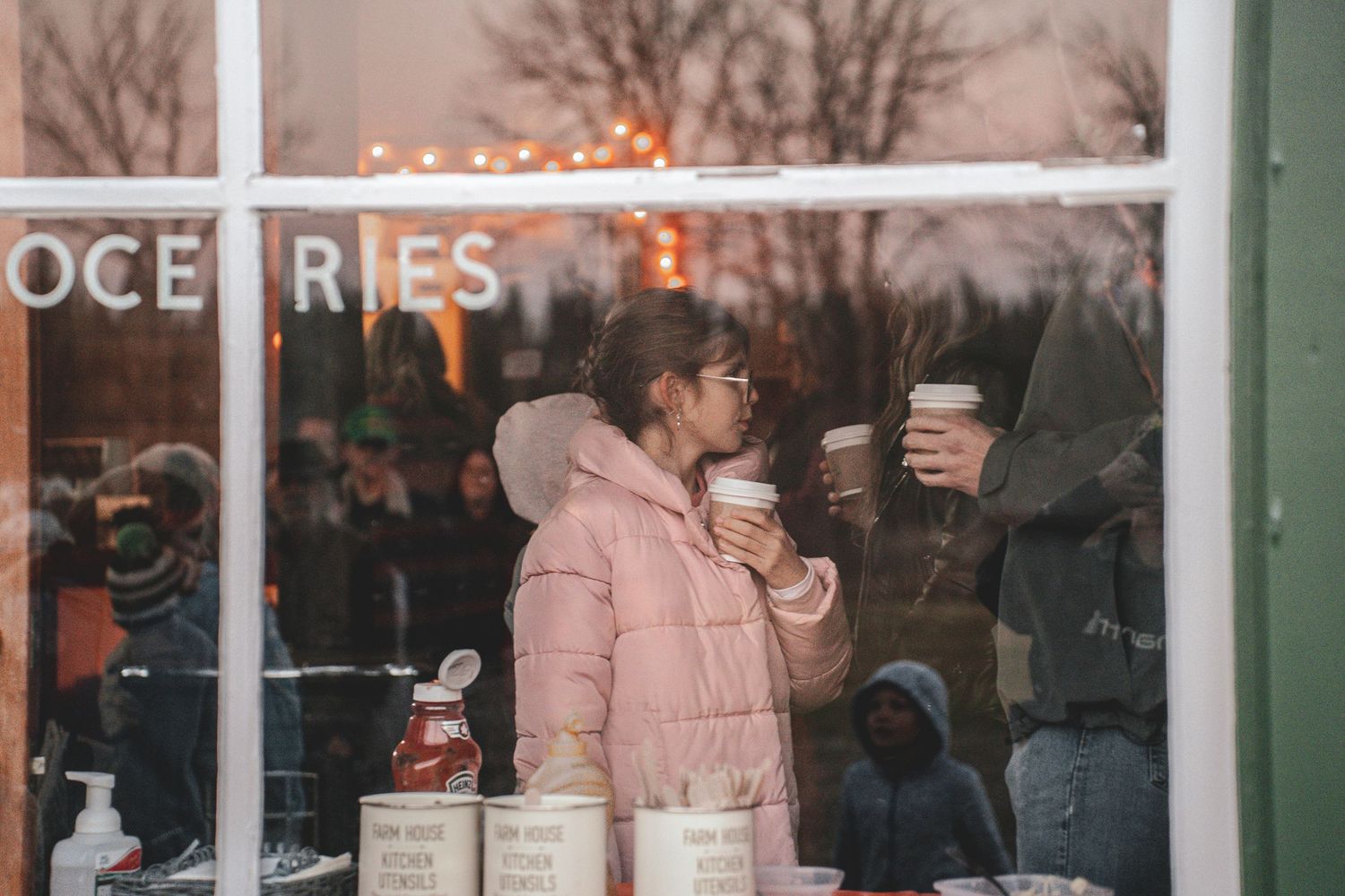 Young woman in pink winter coat holds coffee cups viewed through huble homestead's store on site of their Halloween, commercial event.