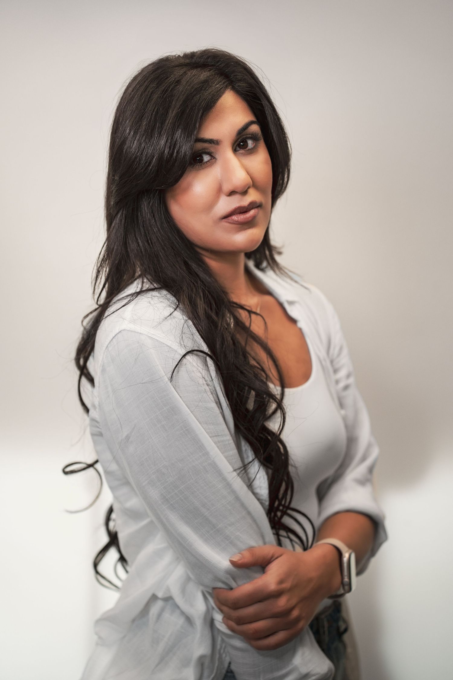 A brunette woman in a white button-up shirt poses sideways against a light gray studio background during her business headshot photoshoot.