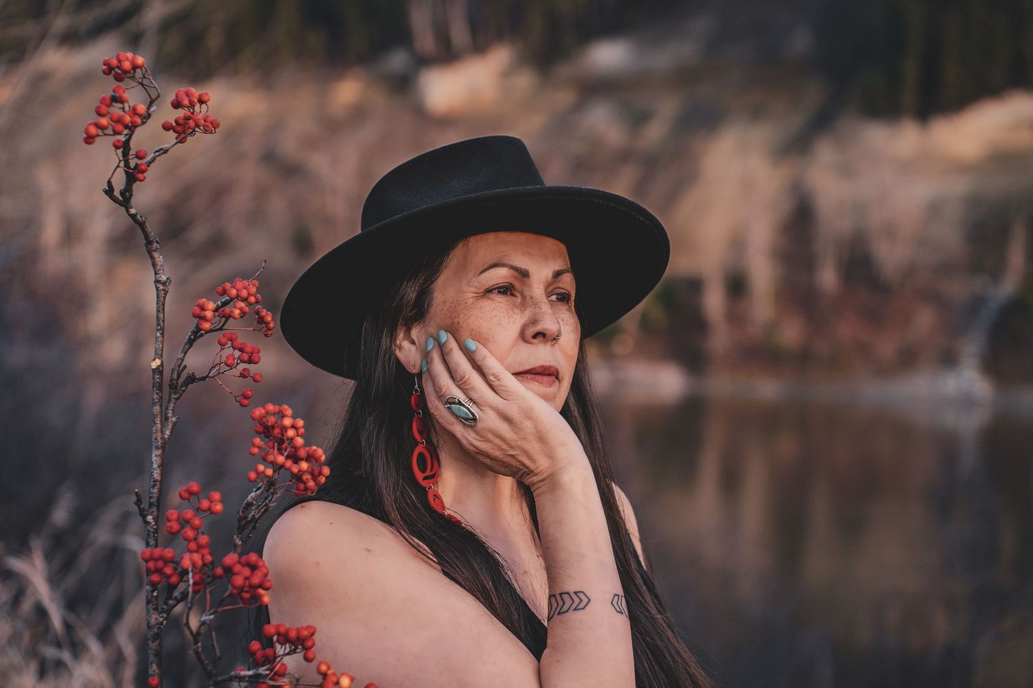 Kym Gouchie in black wide-brim hat with turquoise ring near red berry branches beside a lake during artistic branding photoshoot.
