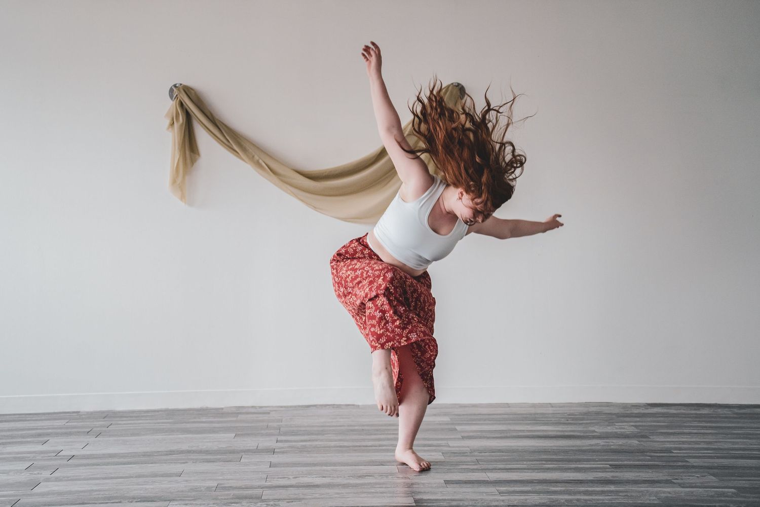 A female dancer leaps gracefully on one foot, her flowing hair and skirt captured mid-motion in a bright studio during branding session.