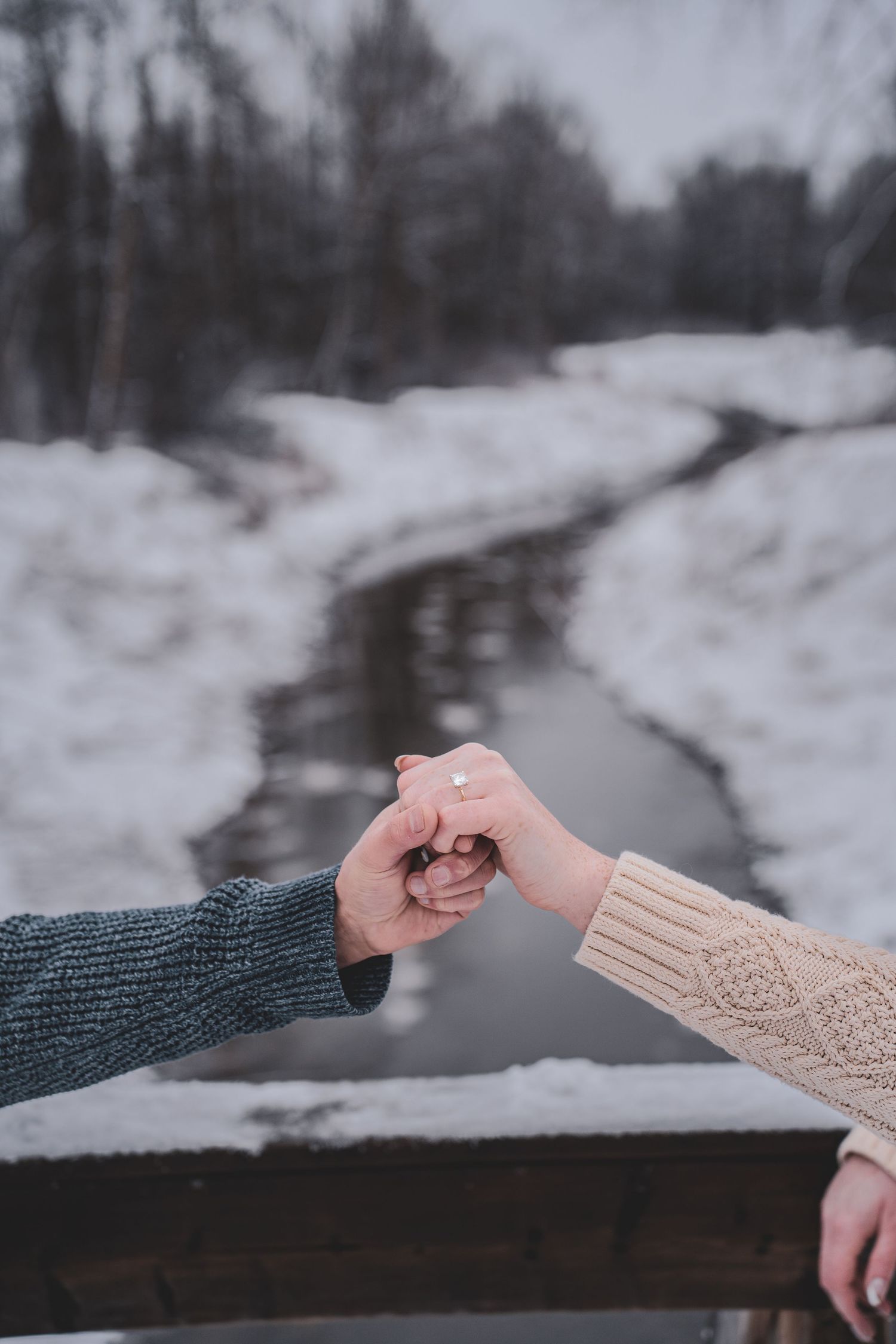 Couple in cozy sweaters make a pinky promise over a snowy winter creek landscape. Capture your engagements in Northern BC