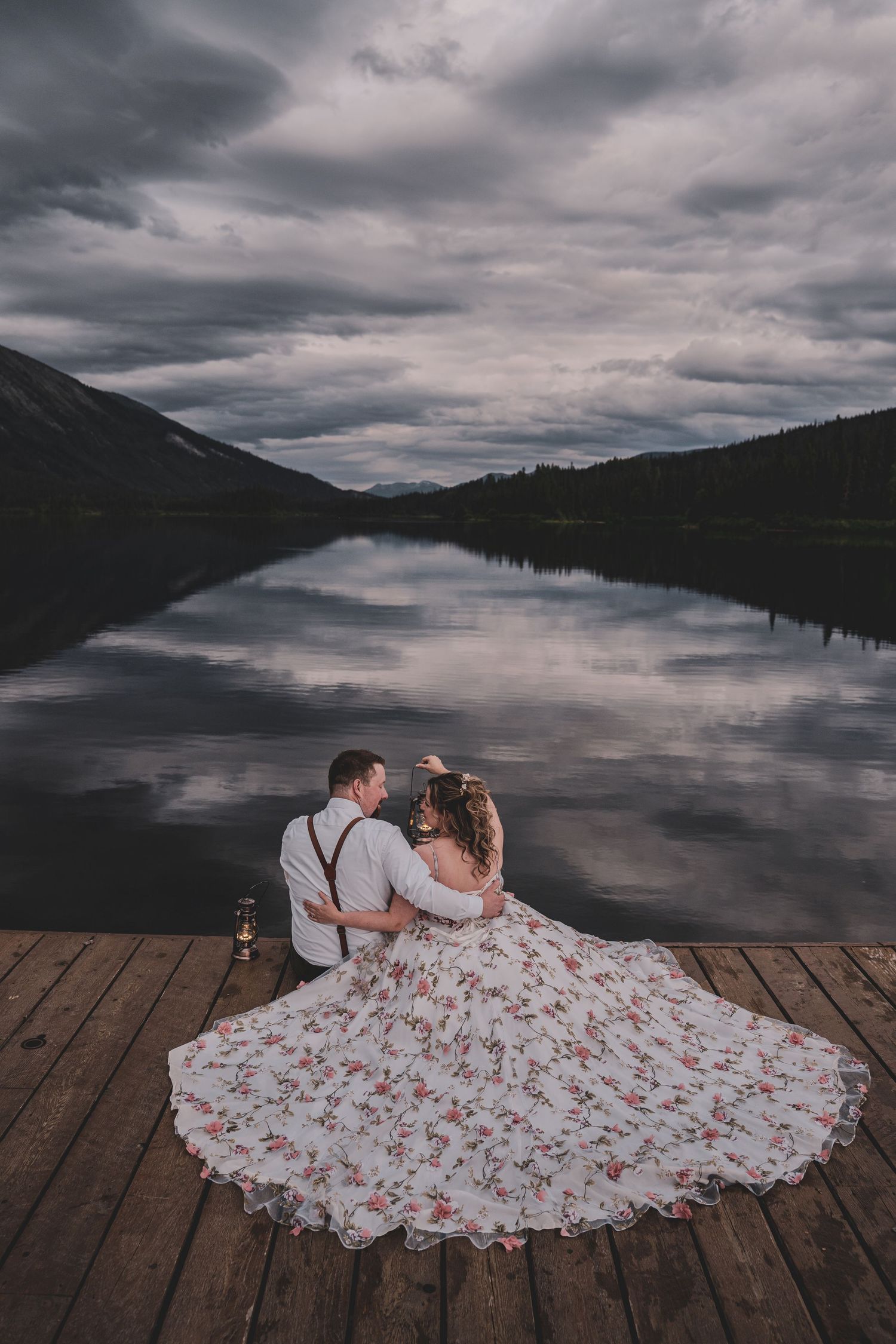 Bride and groom sitting on dock overlooking a reflective mountain lake under dramatic clouds.