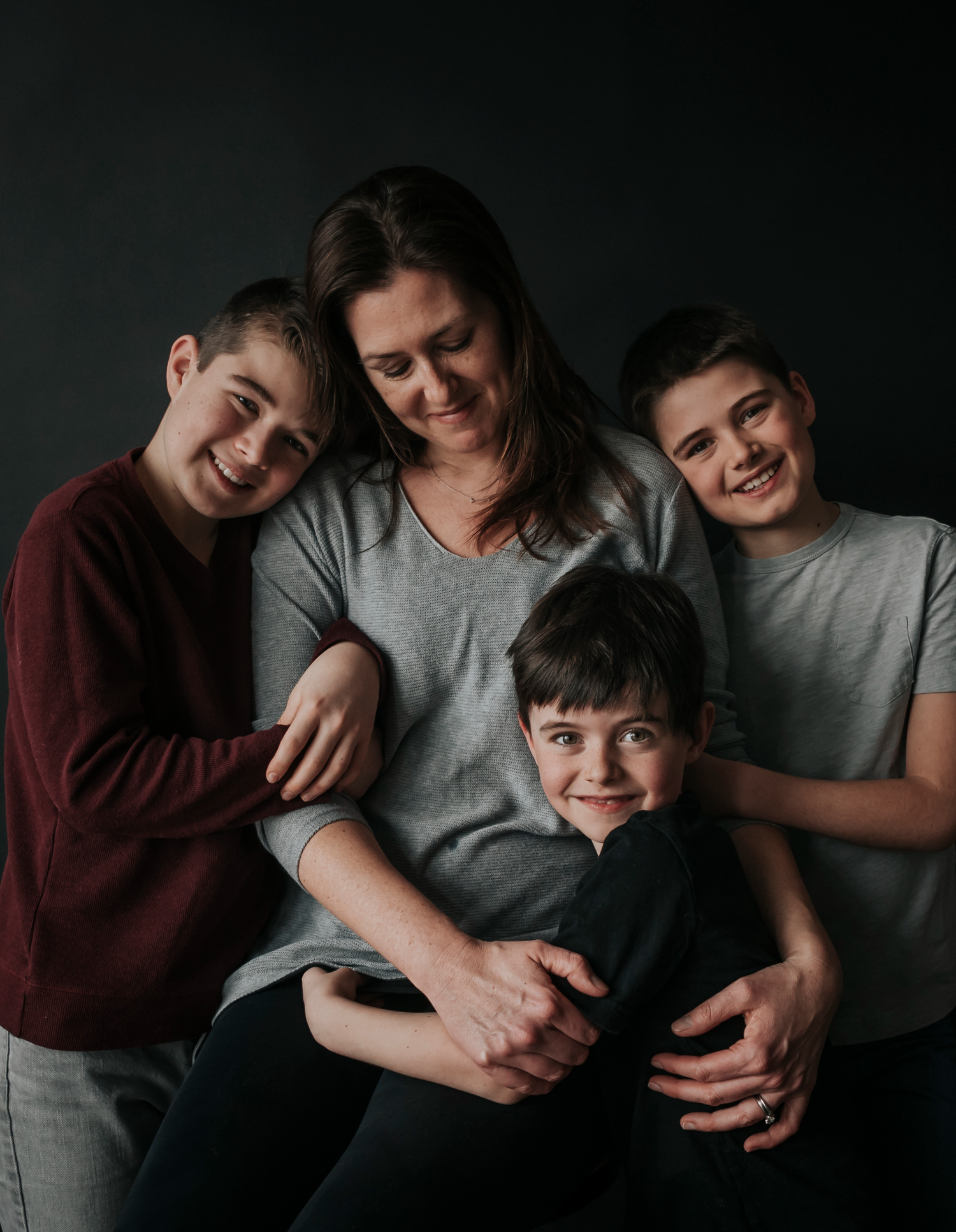 Close-up, moody family portrait of four people embracing tightly against a dark studio background