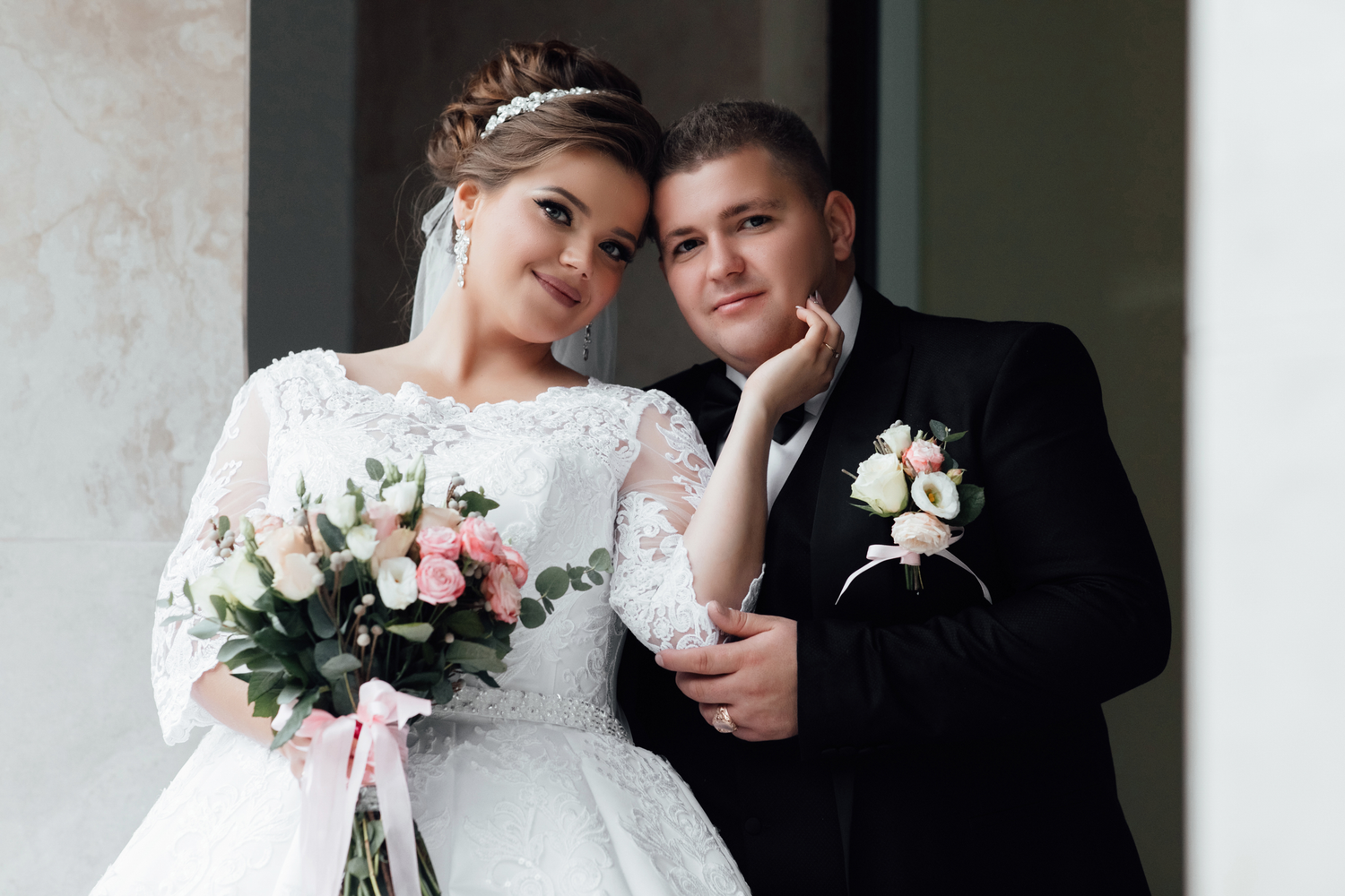 A close-up portrait of a bride and groom posing together. The bride wears a white lace wedding dress and a jeweled headband, holding a bouqu
