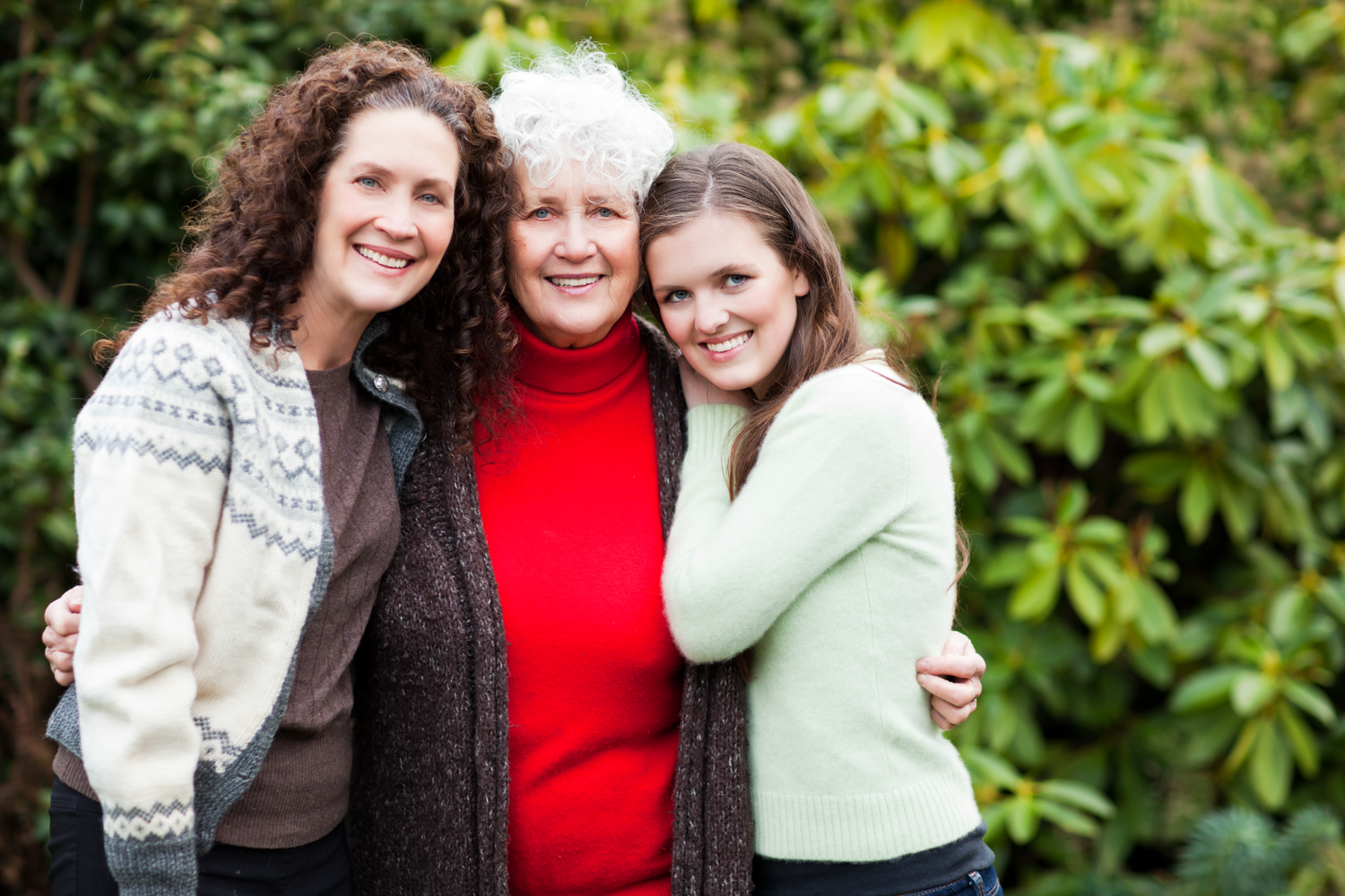 Generational family portrait of three women embracing and smiling outdoors in a lush, green setting