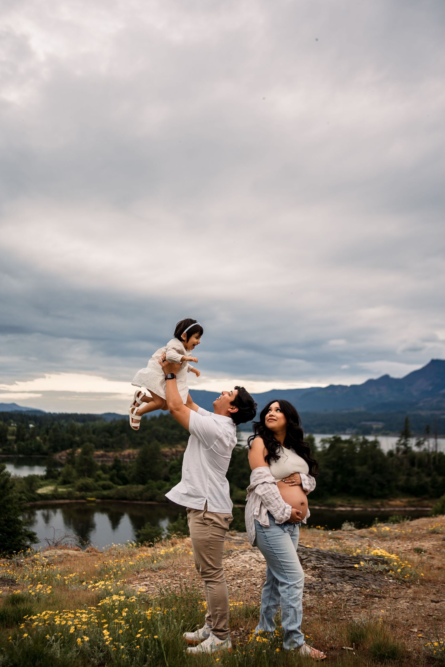 Family playing and lifting child against mountain backdrop with lake view.