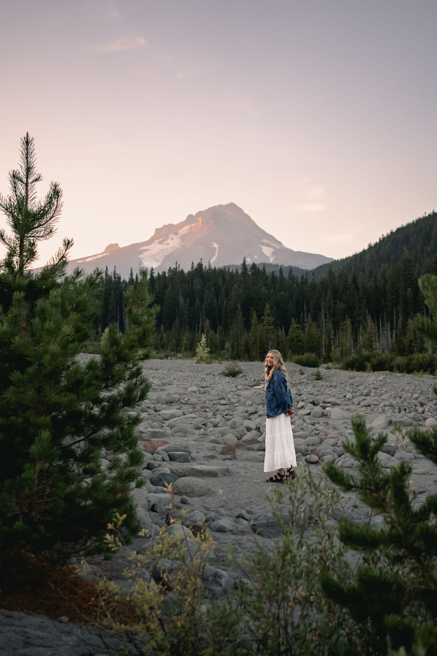 A scenic mountain landscape view with a person in white dress and denim jacket standing on rocky terrain.