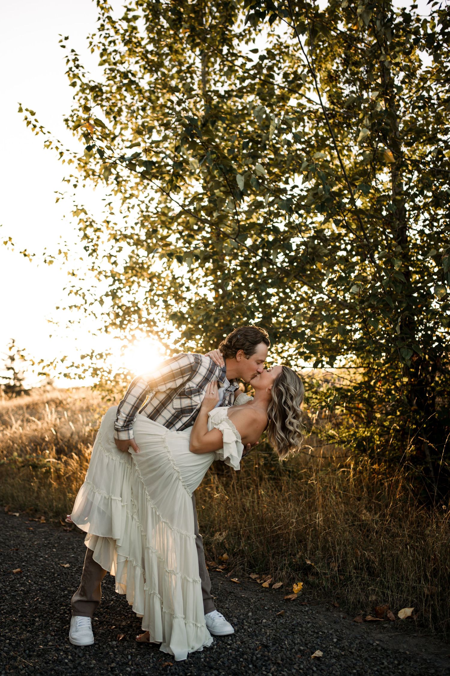 Couple in white outfits dancing together under trees at sunset.