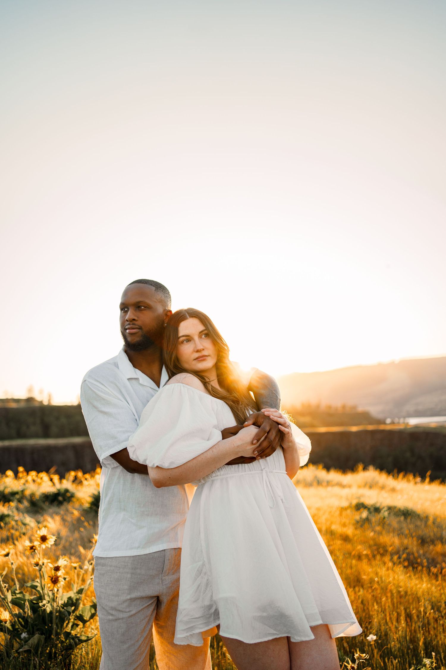 Couple in white outfits embraces in a golden wildflower field at sunset.