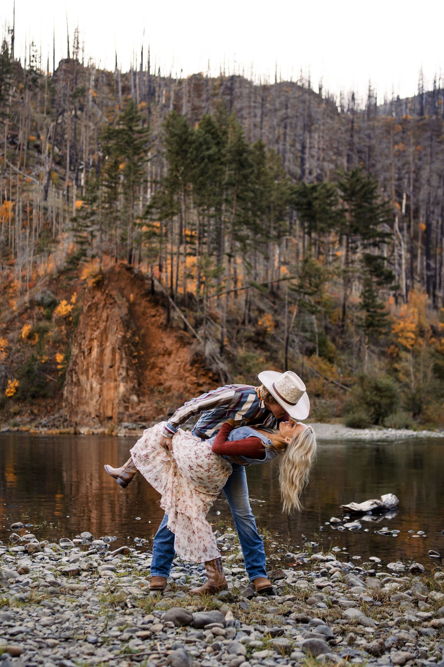 Couple in casual attire posing playfully by a scenic mountain lake.