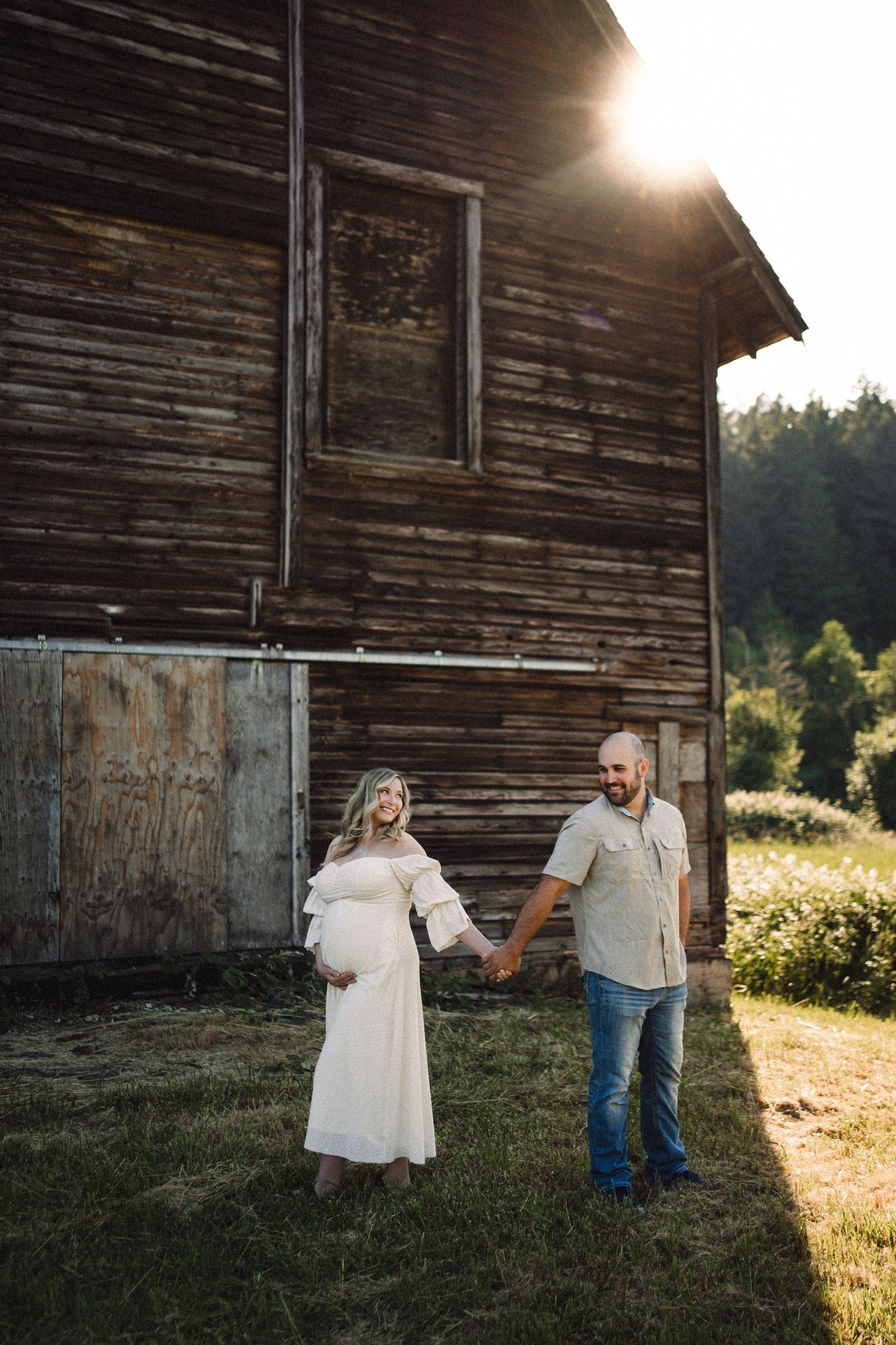 A couple poses together near a weathered wooden barn during golden hour.