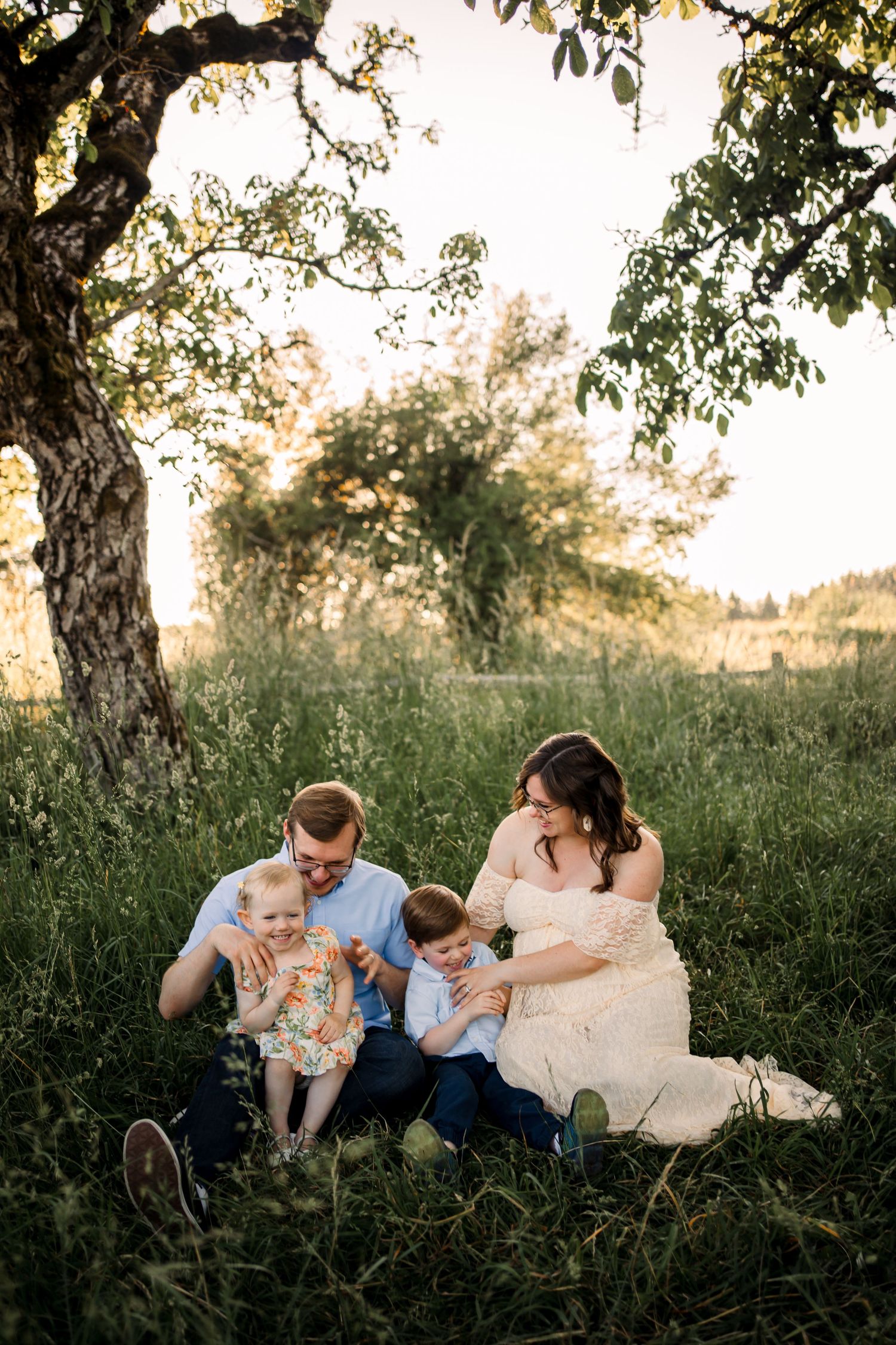 Family relaxing together in a sunlit meadow with tall grass and trees during golden hour.