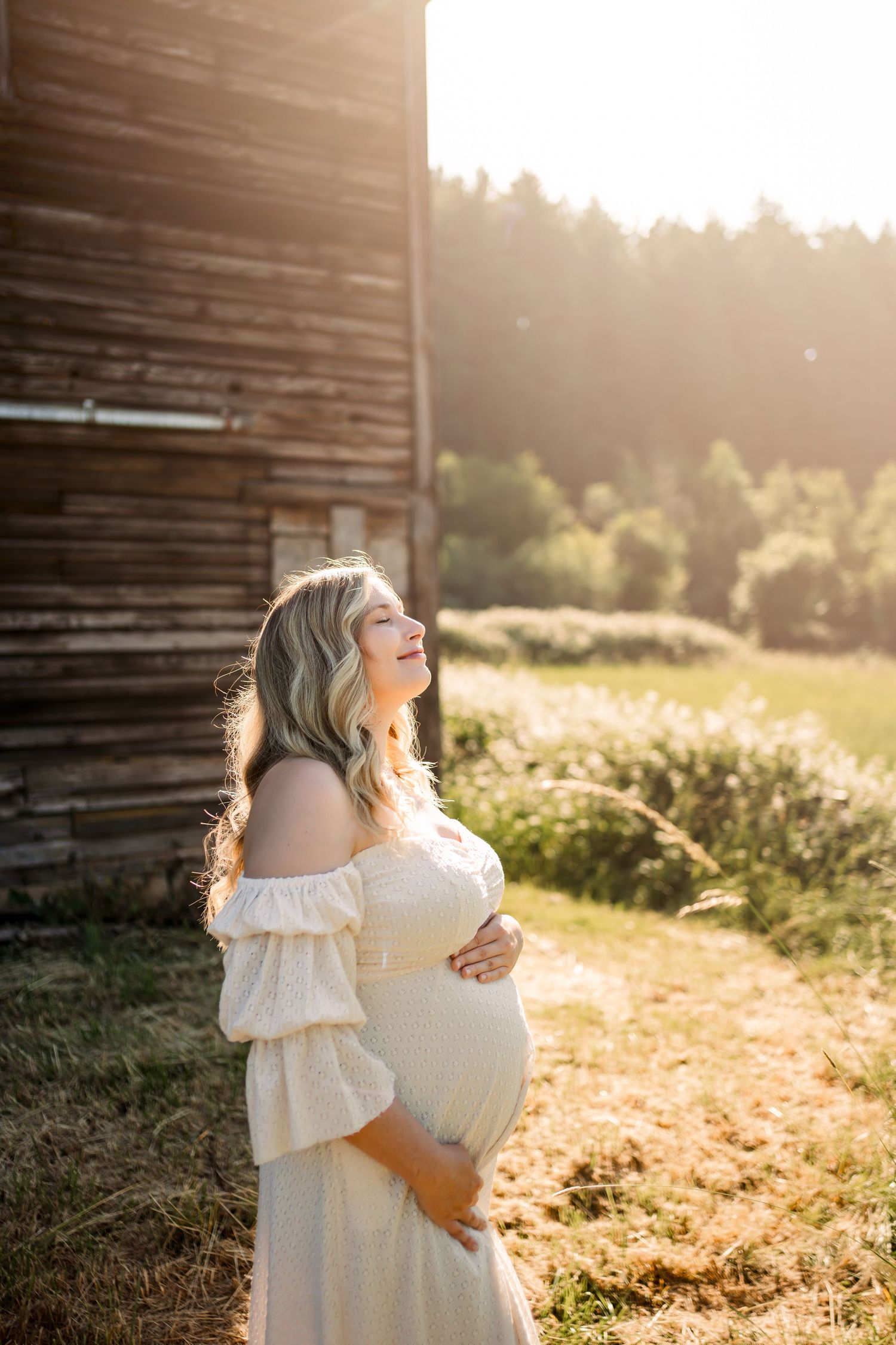 A person in a white dress stands in golden sunlight near a rustic barn.