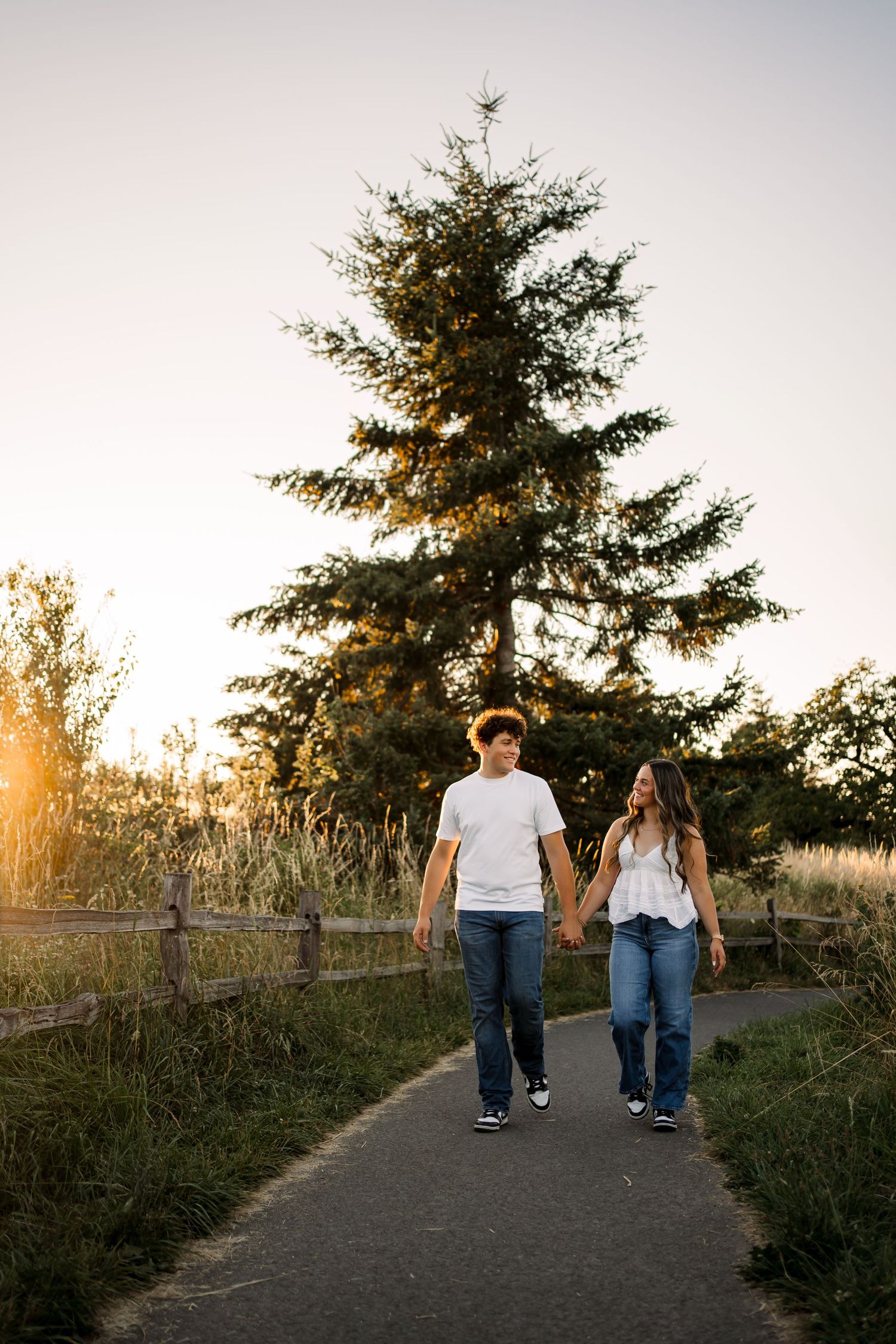 Couple walking hand in hand on a path surrounded by evergreen trees at sunset.