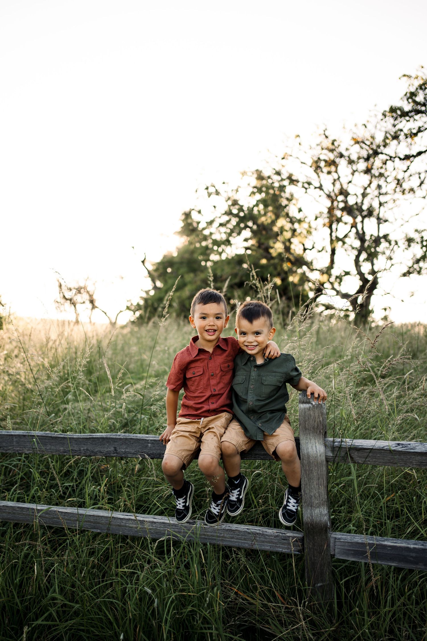 Two children sitting and smiling on a wooden fence at sunset.
