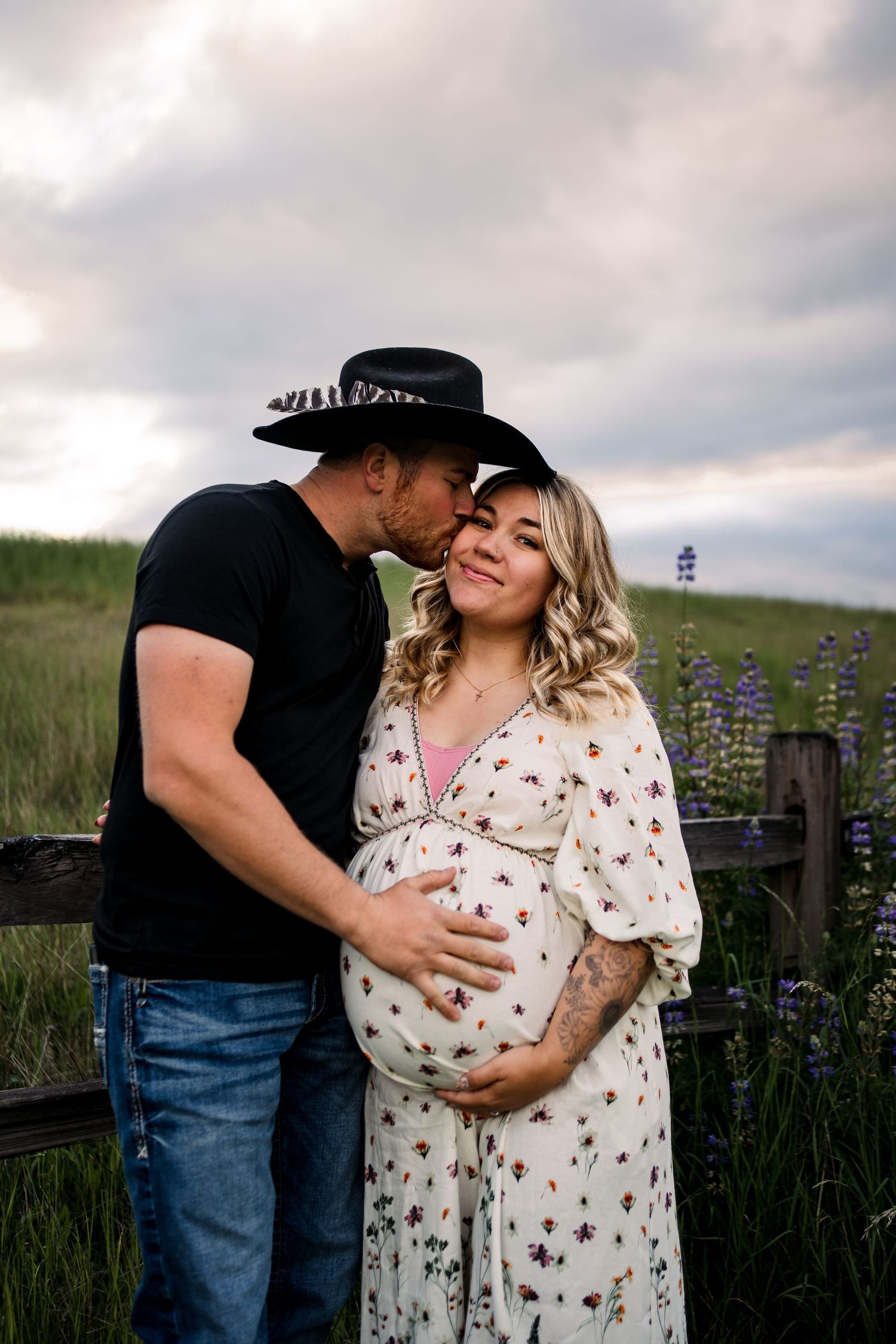A couple embraces in a field of wildflowers during sunset with a rustic fence backdrop.