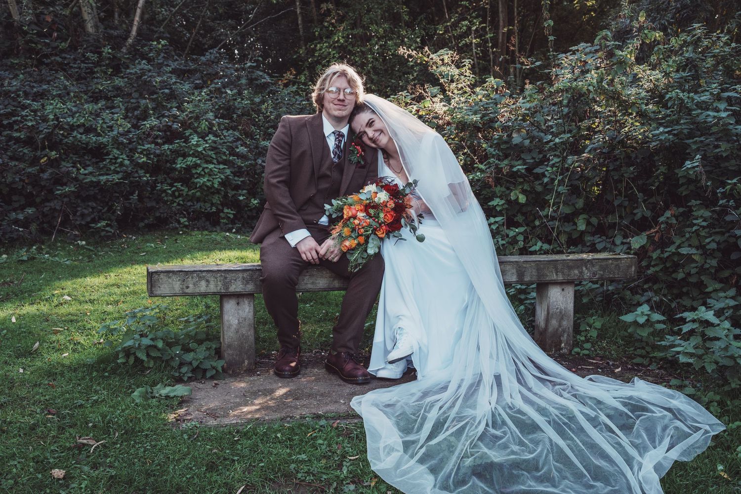 Bride in white gown with veil and groom in brown suit pose on stone bench in lush garden with colorful bouquet.