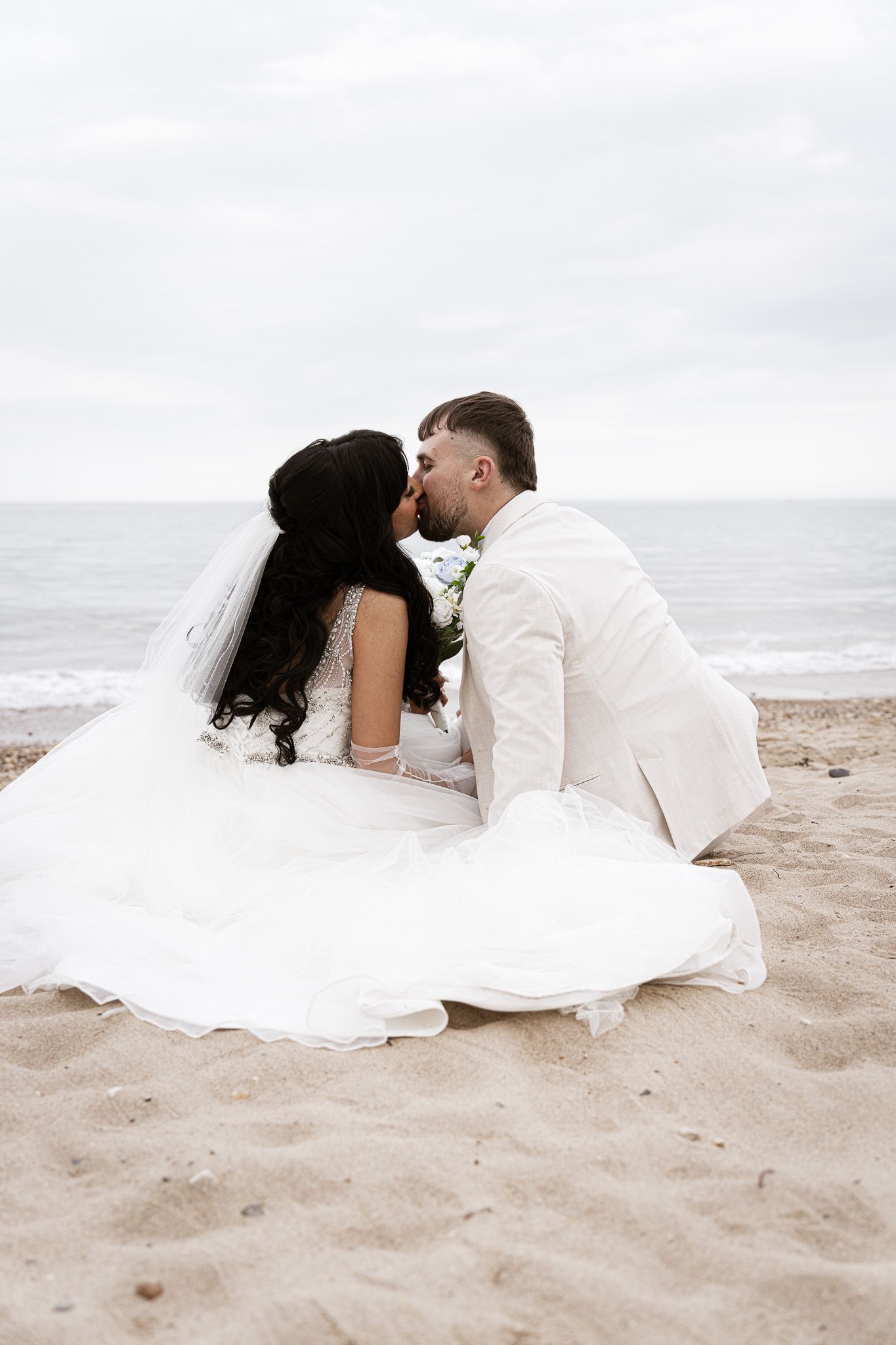 Newlywed couple kissing on sandy beach, bride in white gown with veil, groom in white suit, ocean backdrop.