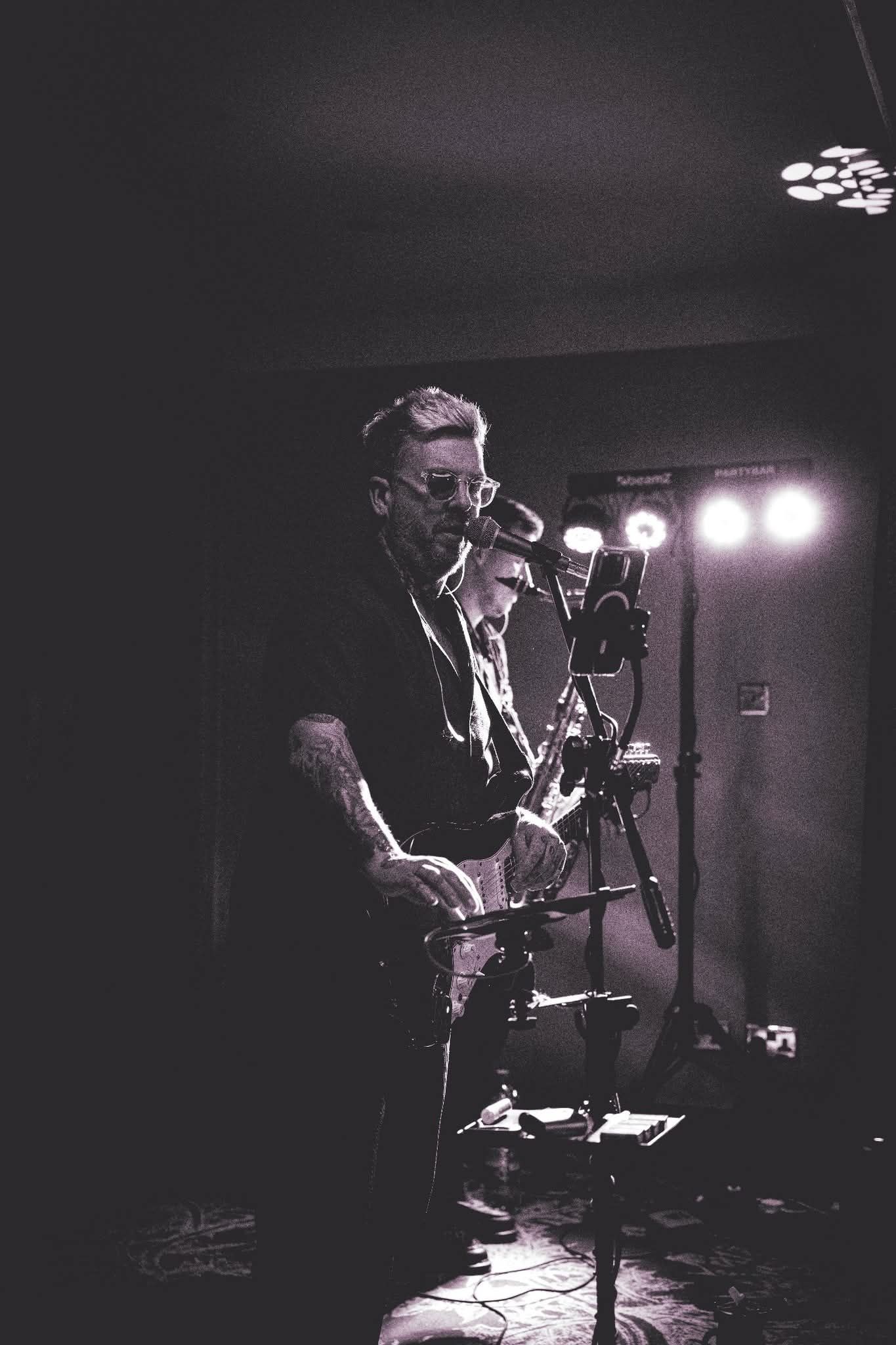 Musicians perform in a dimly lit venue with dramatic stage lighting creating moody black and white ambiance.