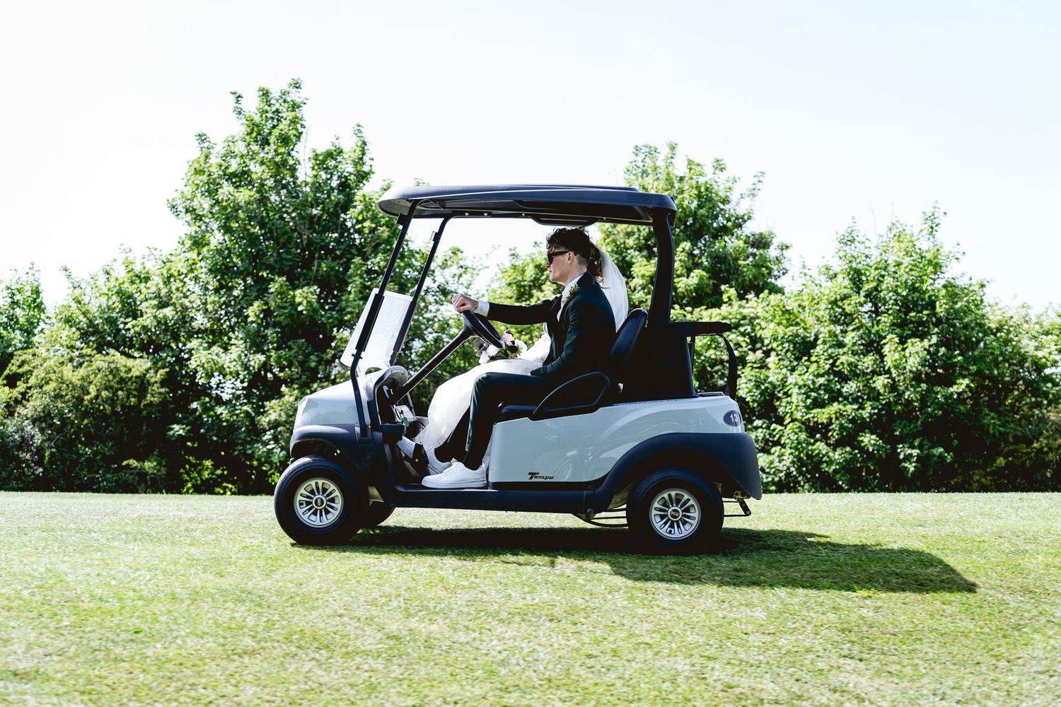 Person in formal attire driving a white golf cart on a lush green golf course on a sunny day.