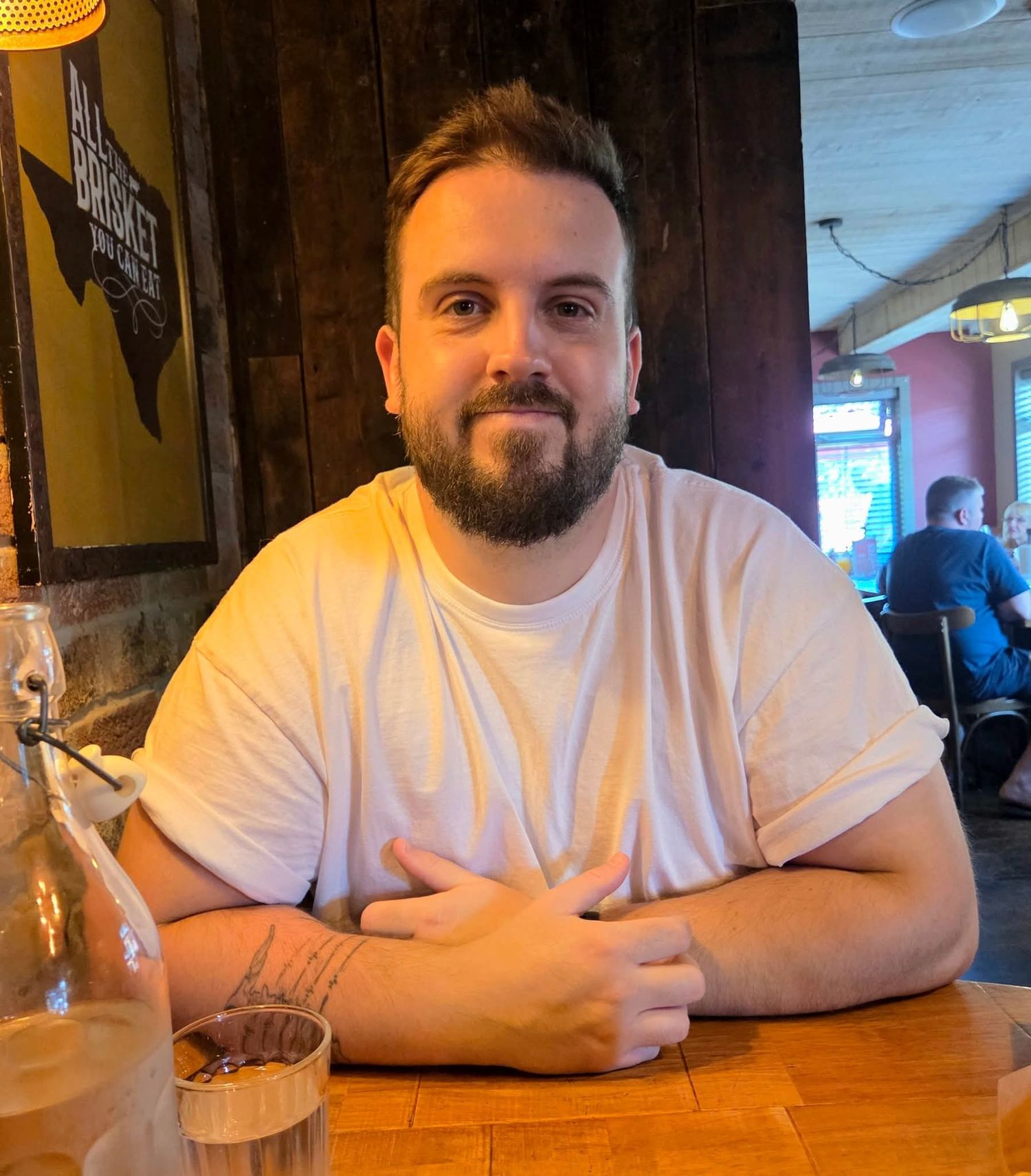A person in a white t-shirt with a beard sits at a wooden bar table with a drink in a casual restaurant setting.