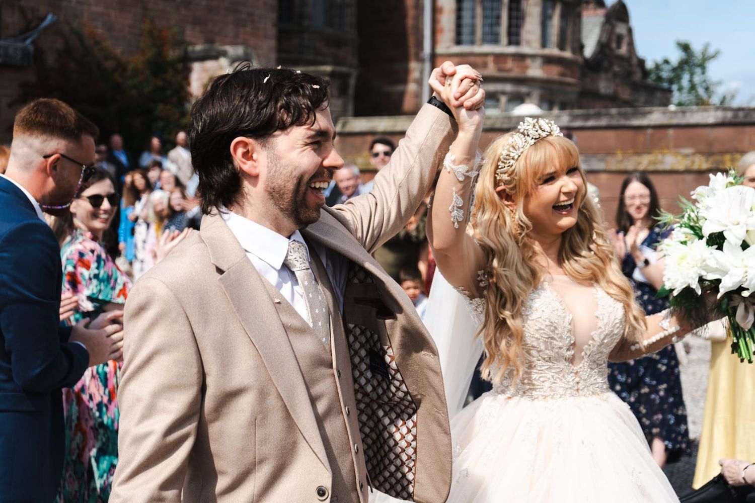 Wedding guests celebrate and embrace during an outdoor reception with bright sunlight and floral decorations.
