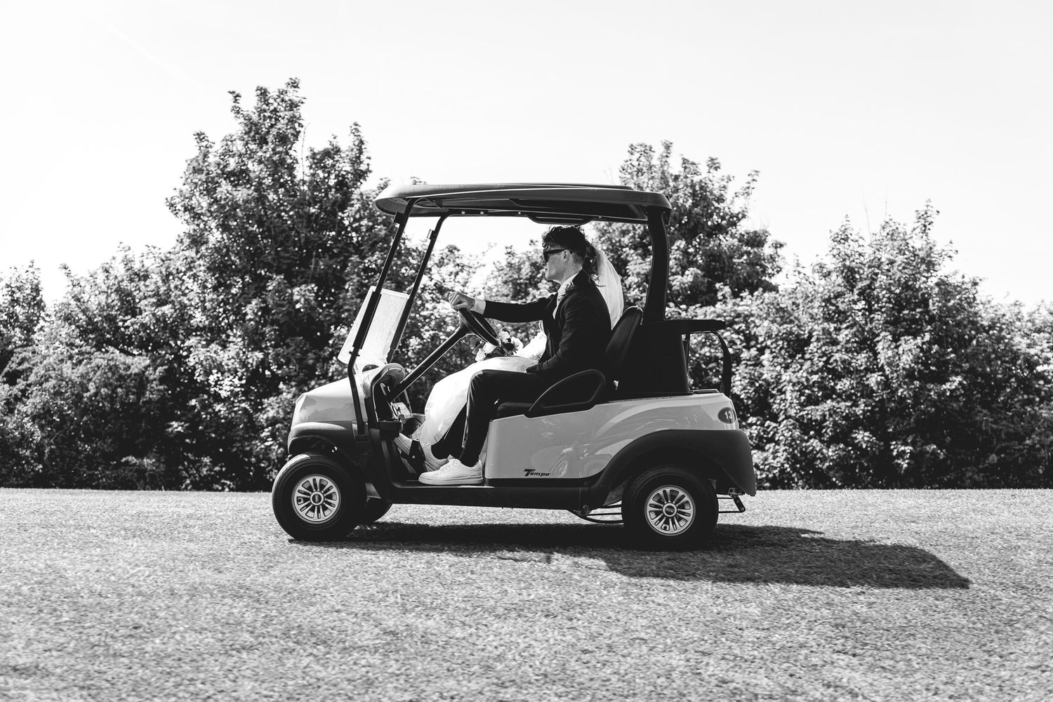 Two people in formal attire ride a golf cart on a sunny day, surrounded by lush trees, in a black and white photo.