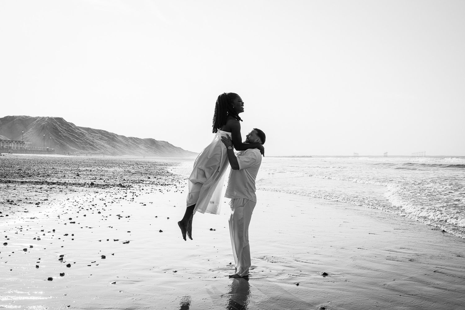 Black and white photo of a couple embracing on a beach, with ocean waves and mountains in the background.