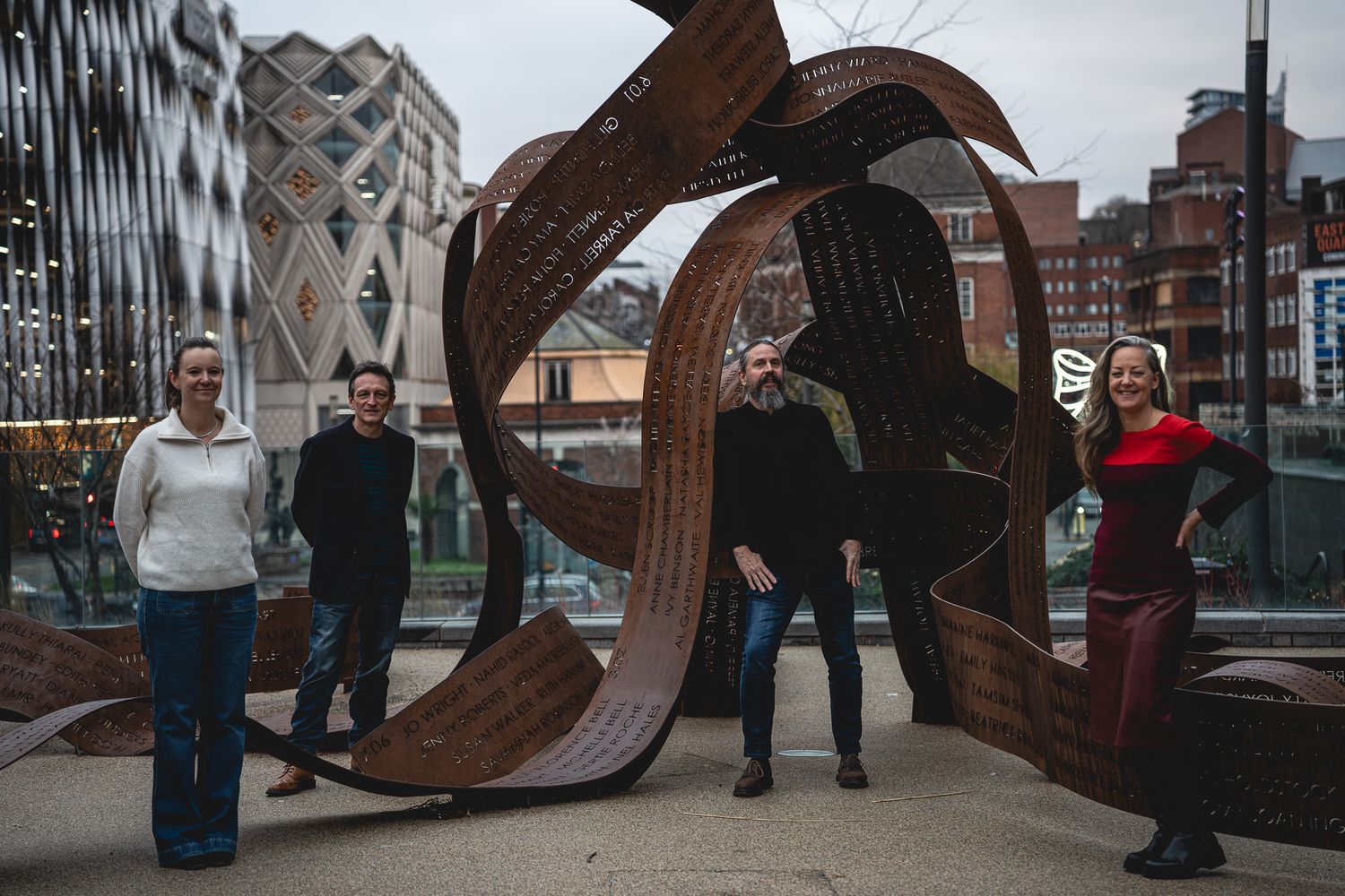 People pose with a large curved metal sculpture on a bridge in an urban setting.