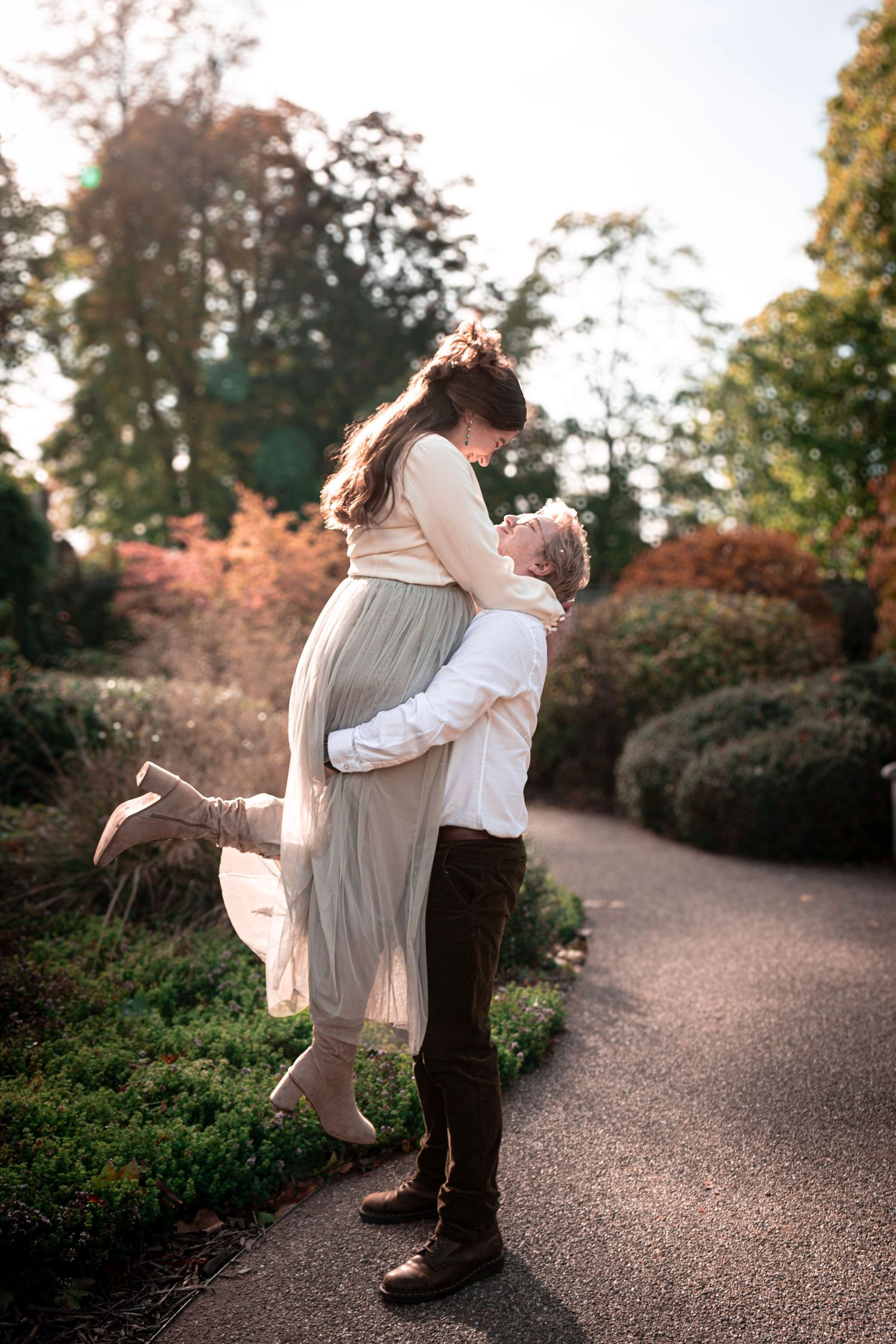 A romantic couple embraces on a garden path during sunset, with fall foliage and trees in the background.
