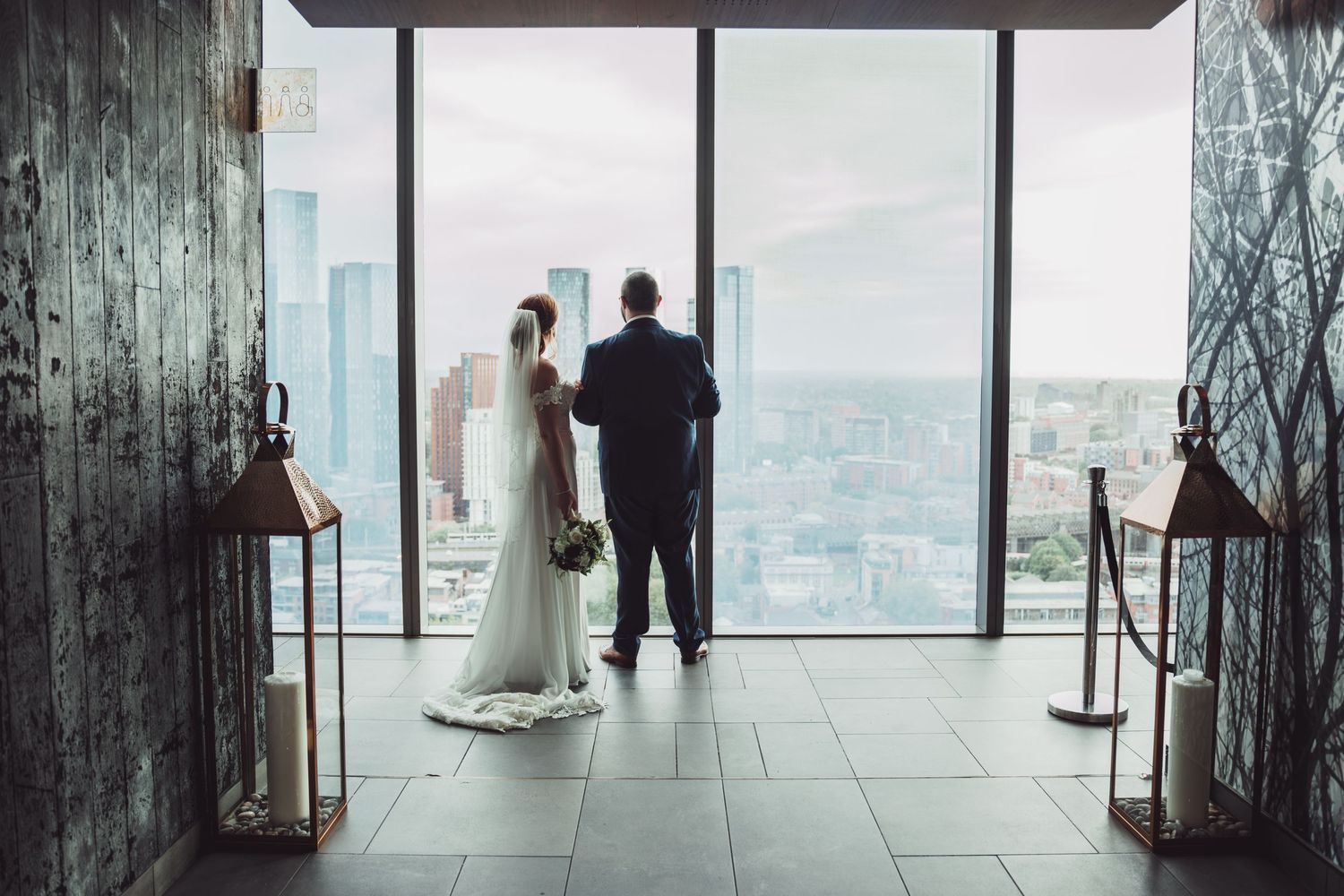 A bride and groom stand before floor-to-ceiling windows overlooking a city skyline in an elegant wedding venue.