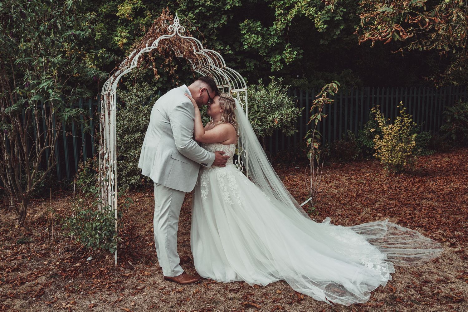 A bride and groom share a kiss under a white garden arch surrounded by lush greenery on their wedding day.