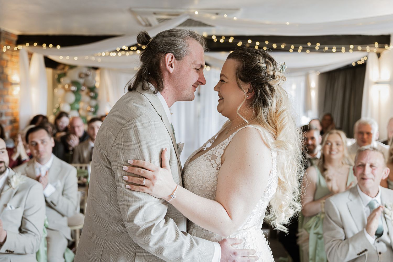 A bride and groom share their first dance, smiling at each other in a warmly lit wedding reception venue.