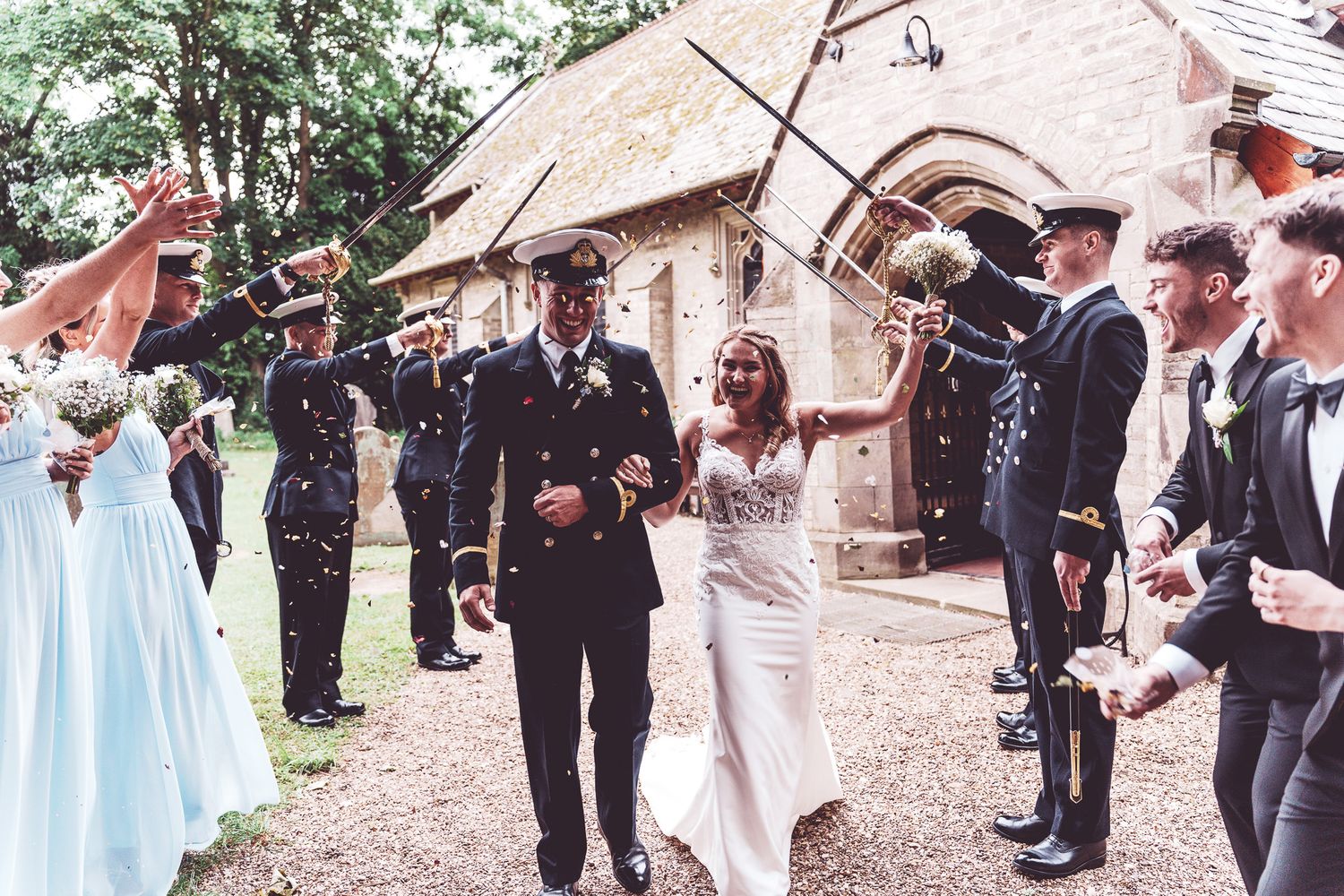Military wedding couple exits church under swords arch with confetti, bride in white gown, groom in uniform.