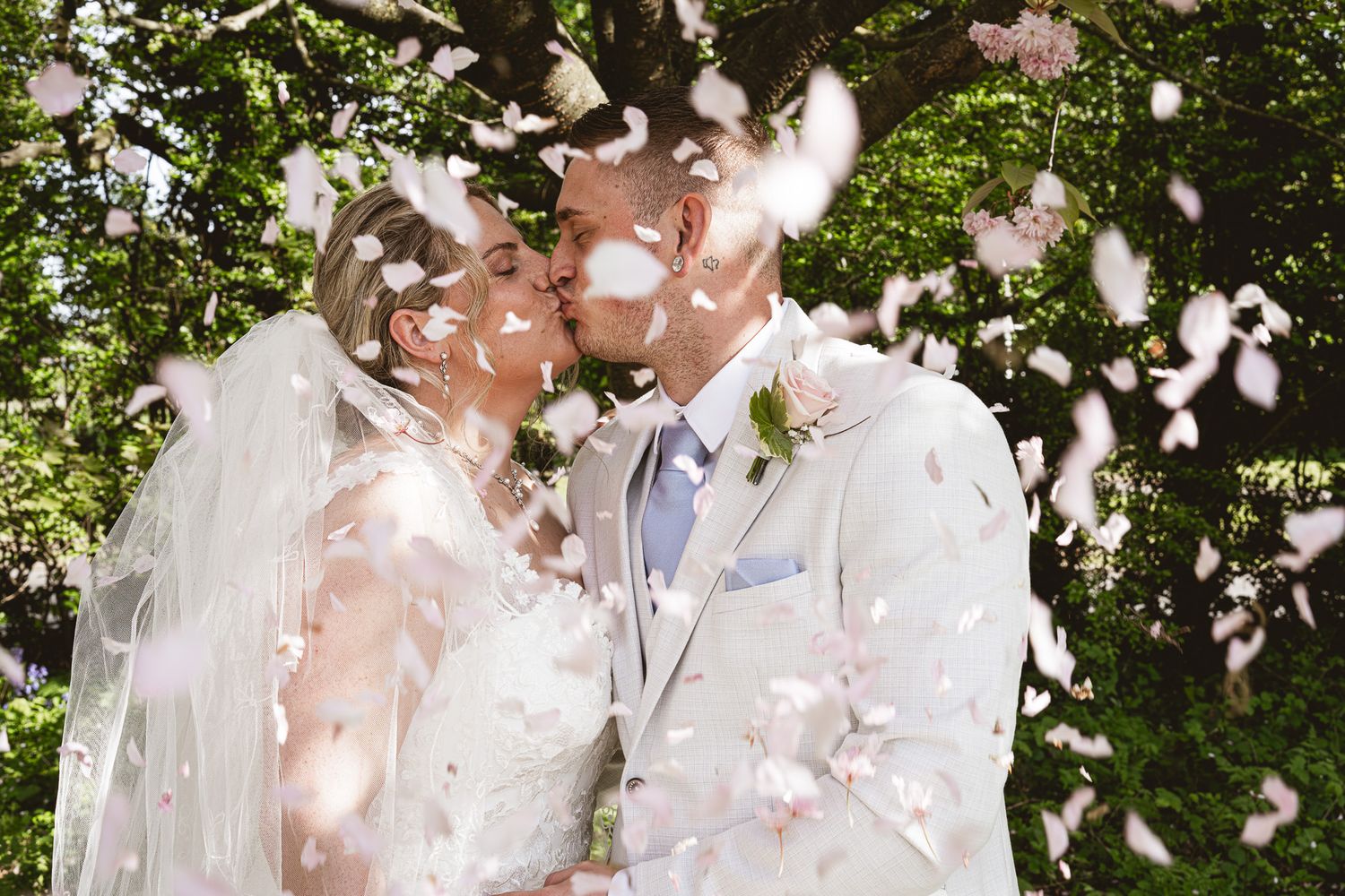 Newlywed couple sharing a romantic kiss under a shower of flower petals outdoors on their wedding day.