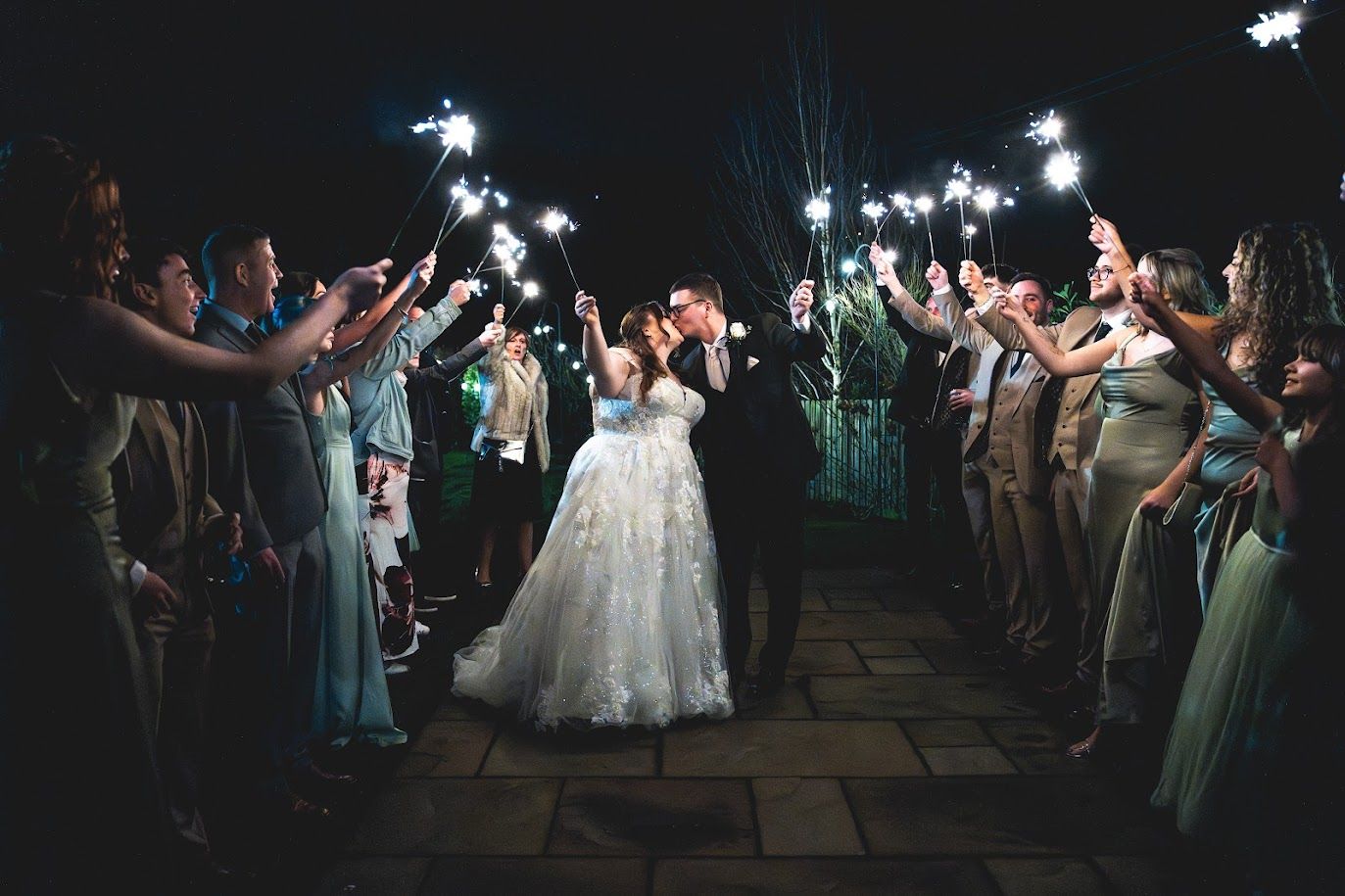 Wedding guests dance and celebrate during an evening reception at a rustic outdoor venue under string lights.